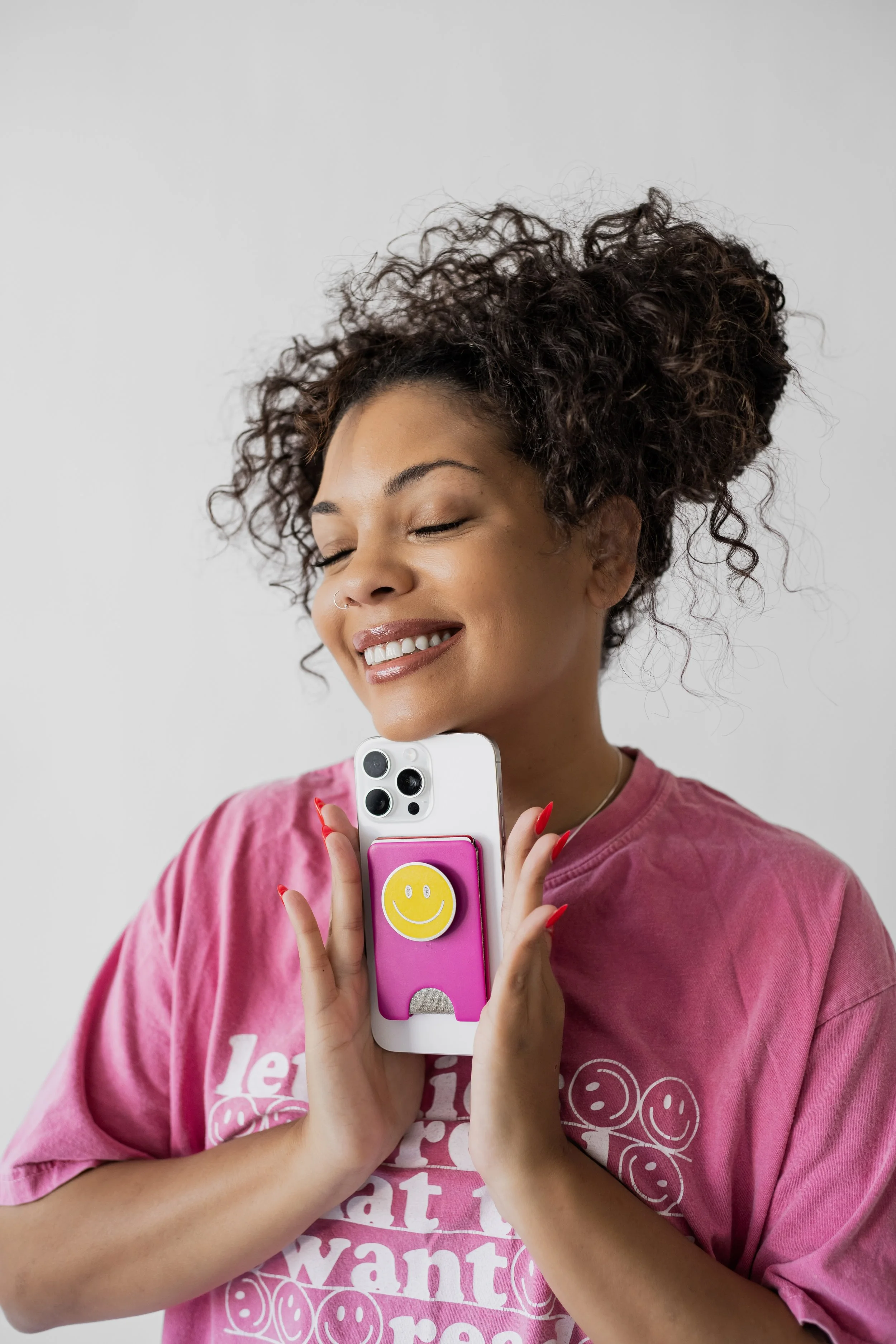 A woman with curly hair smiling with her eyes closed, holding a smartphone with a pink pop socket and a yellow smiley face sticker, wearing a pink T-shirt with white text and smiley faces.