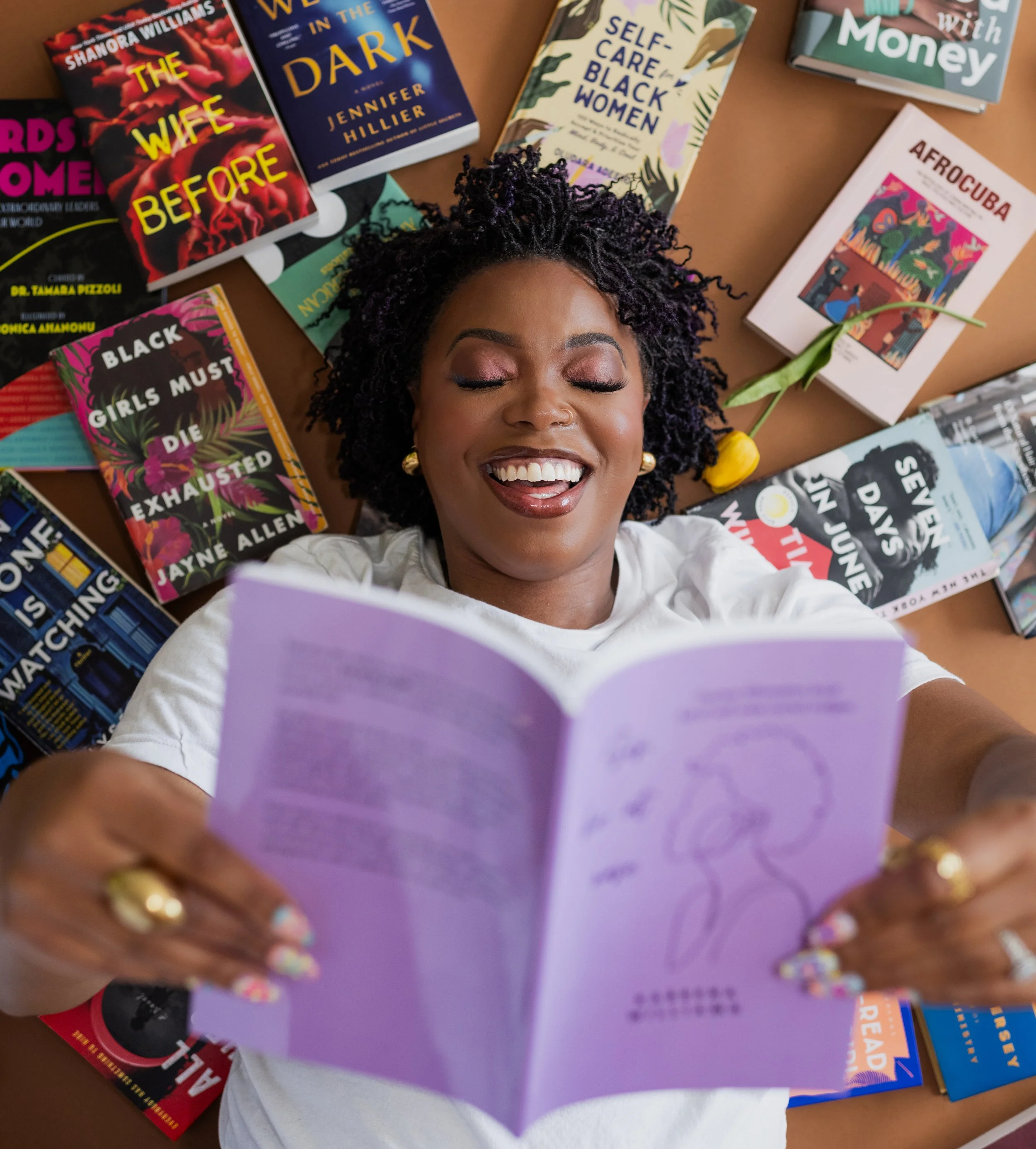 A woman with curly hair and gold earrings lying down with her eyes closed, smiling while holding an open purple book. Several other books are spread around her on a brown surface, including titles about Black women, self-care, and social justice.