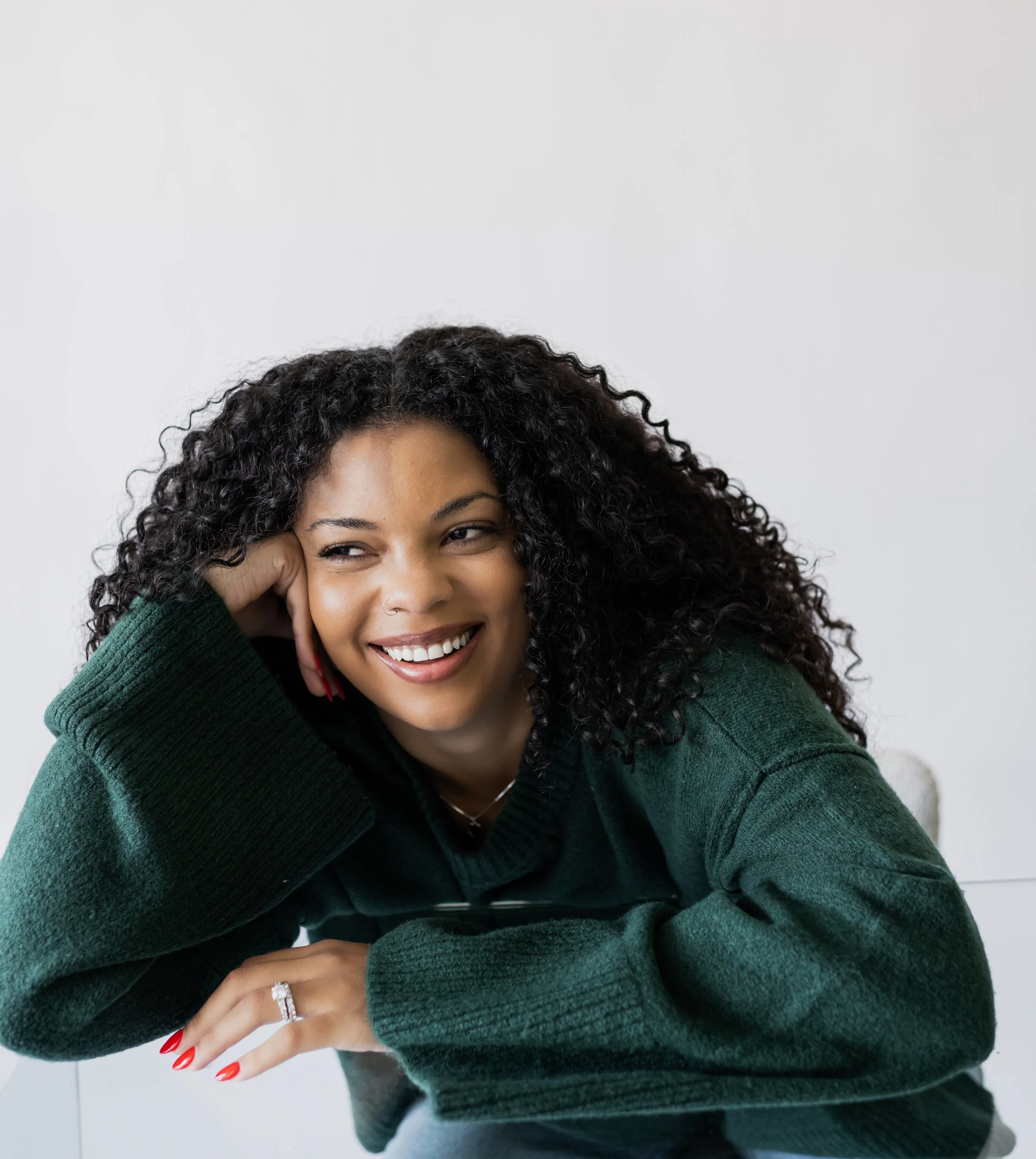A woman with curly black hair smiling and resting her chin on her hand, sitting in front of a plain white wall