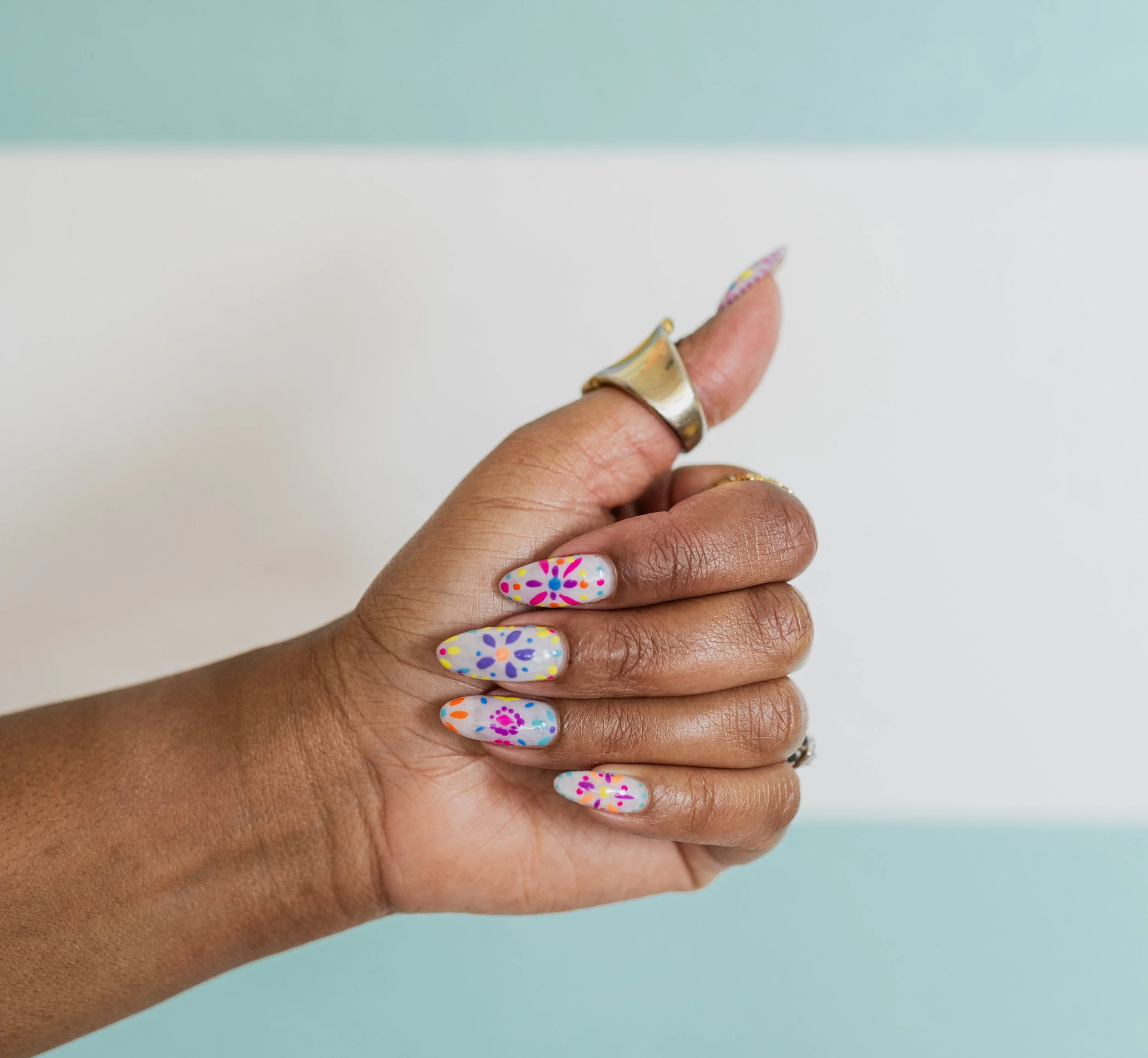 A person's hand with colorful floral patterned nail art, jewelry including rings, against a split pastel blue and white background.