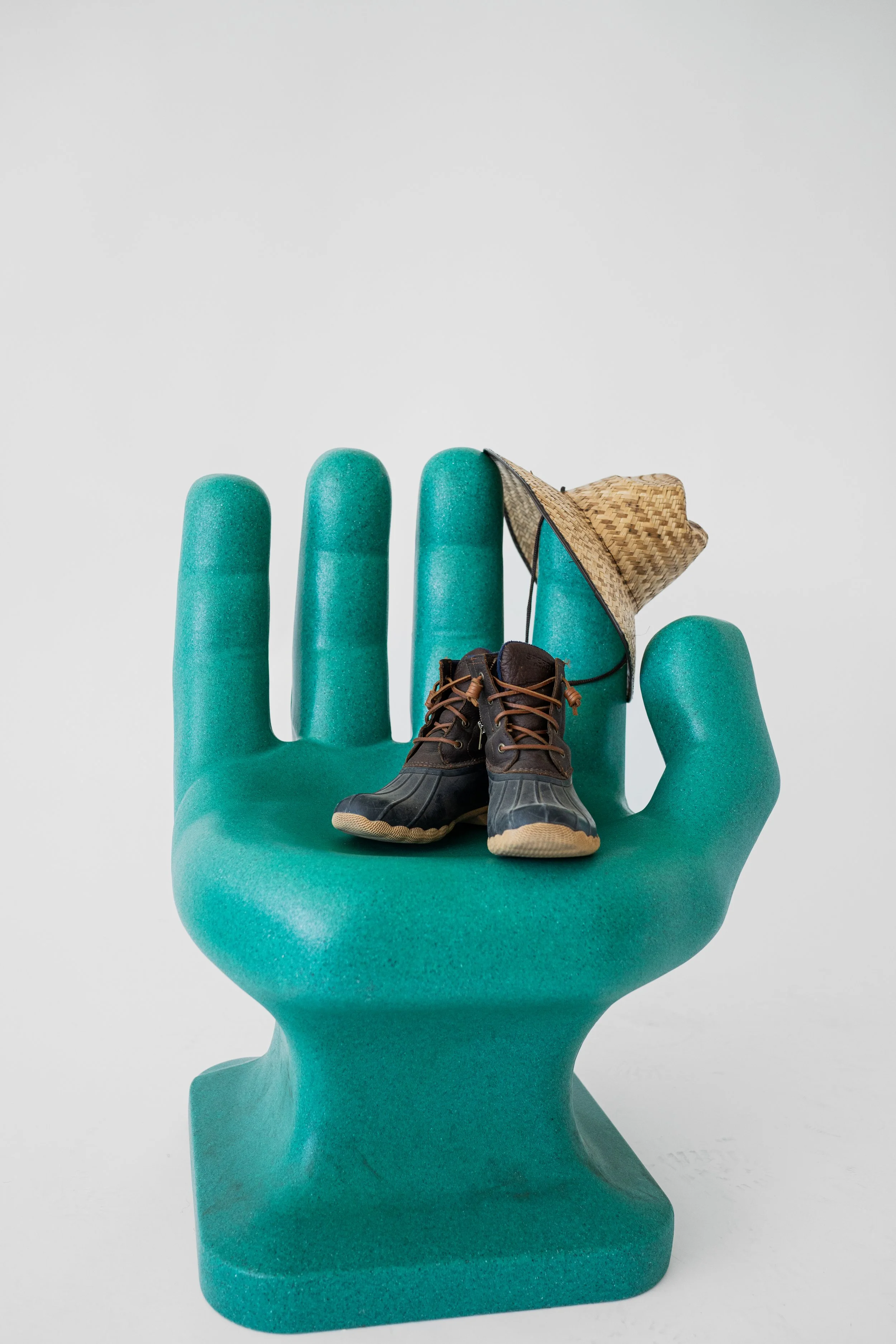 A teal-colored hand-shaped sculpture holding miniature work boots, a straw hat, and a small woven bag.