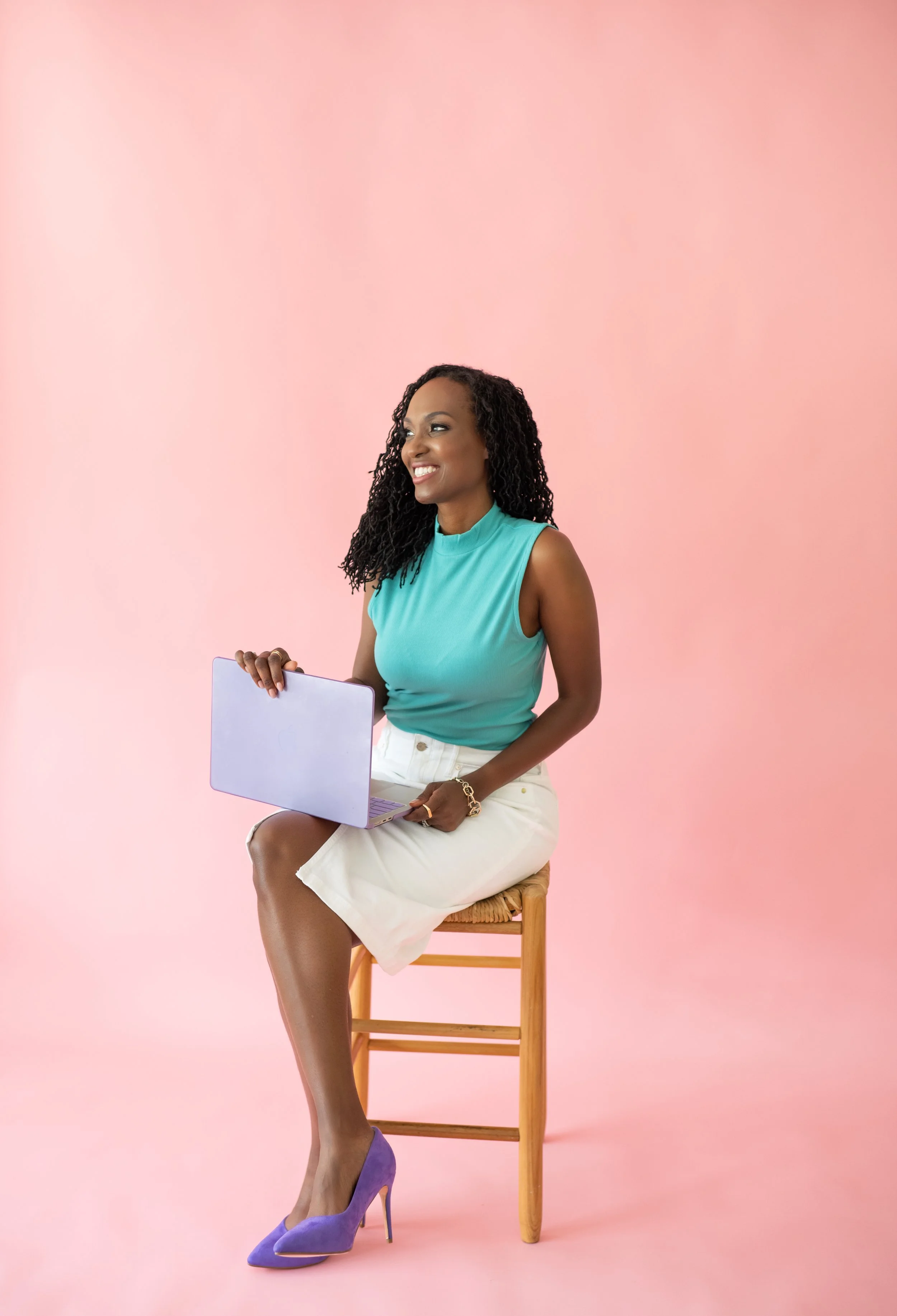 A woman with curly black hair sitting on a wooden chair against a pink background, holding a purple laptop, smiling, wearing a turquoise sleeveless top, white skirt, and purple high heels.