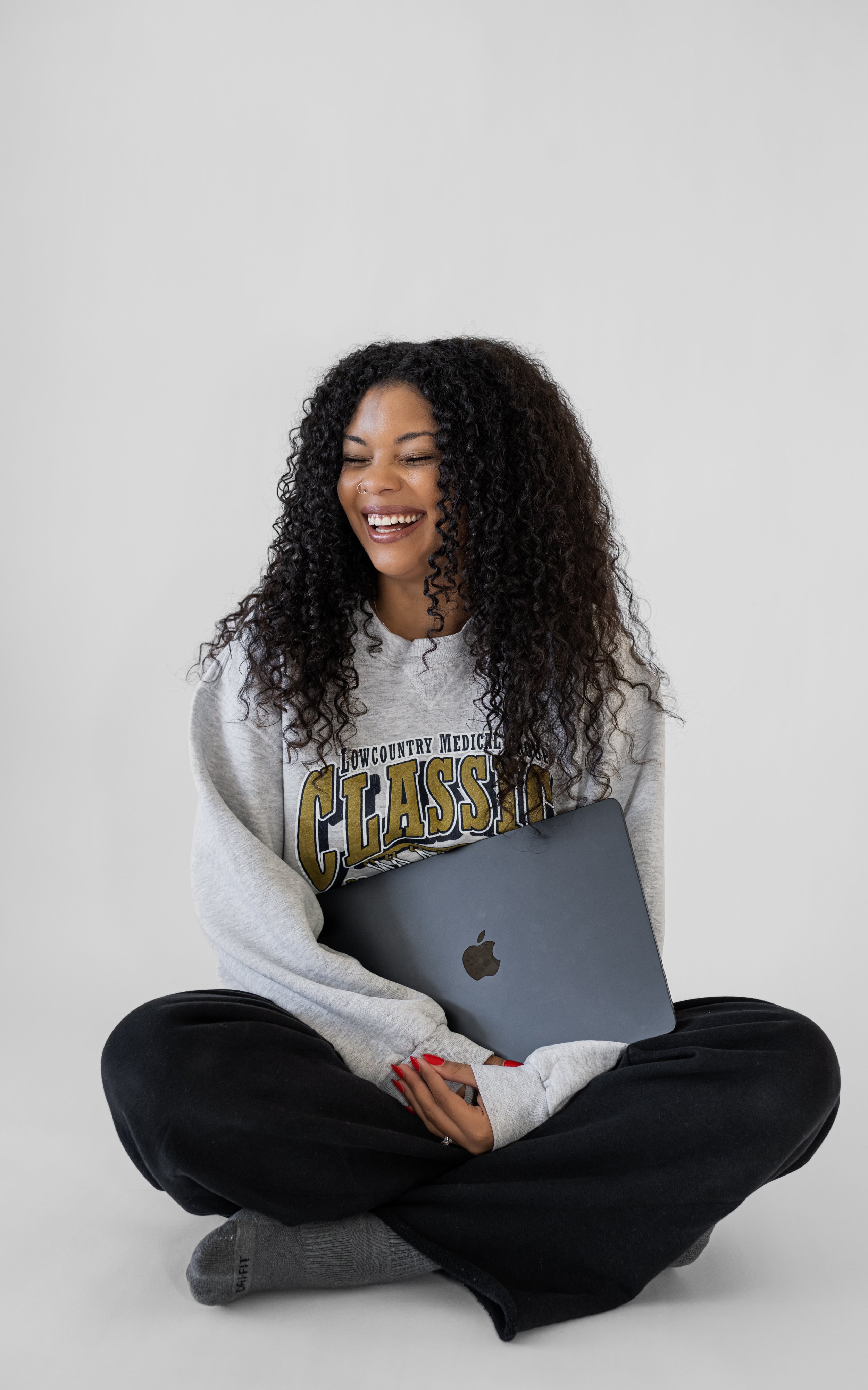 A young woman sitting cross-legged, holding a silver MacBook, smiling with eyes closed, wearing a gray sweatshirt with 'Lowcountry Medical Classic' printed on it, black pants, and gray socks, against a plain gray background.