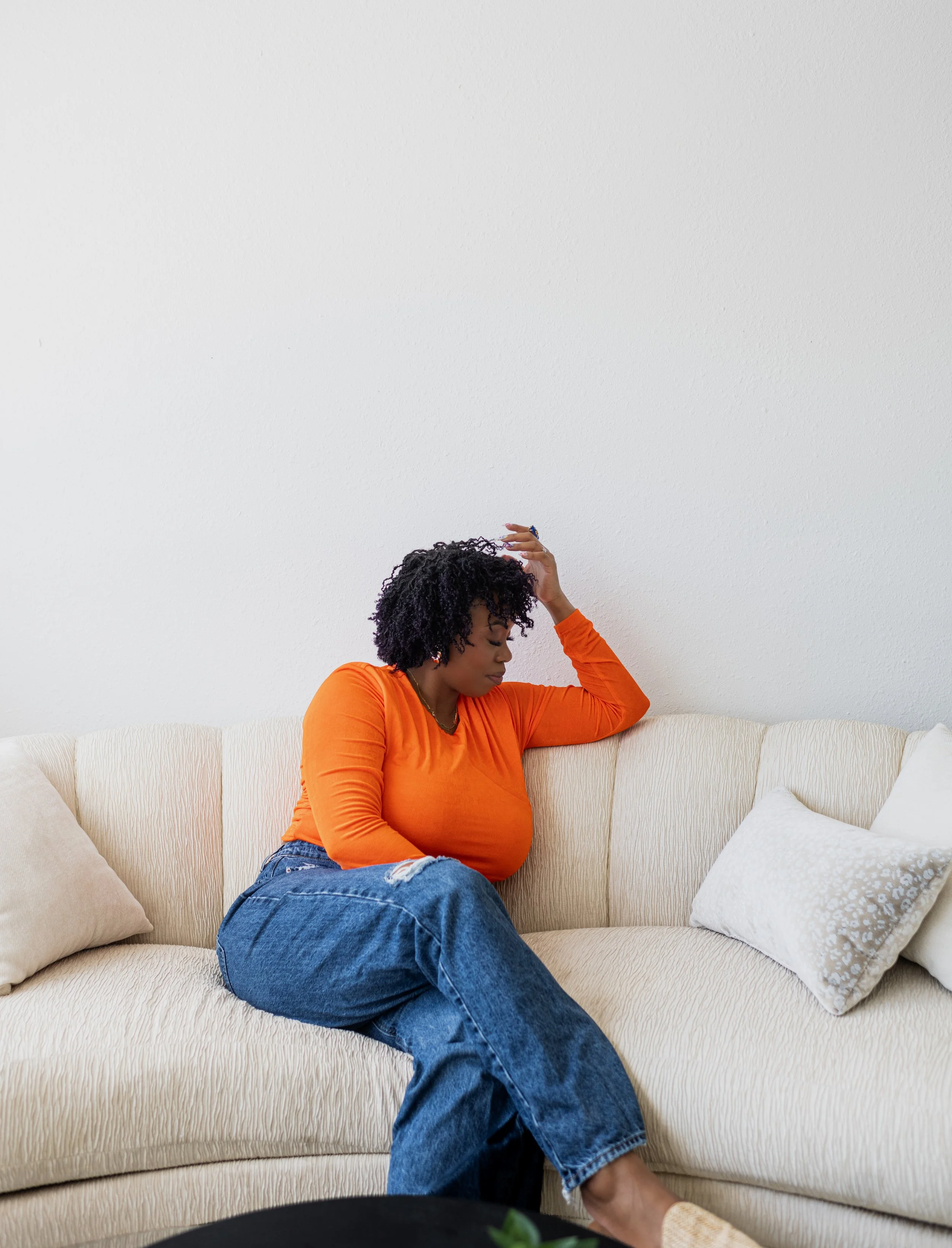 A woman with curly black hair, wearing an orange long sleeve shirt and blue jeans, sitting on a cream-colored sofa with white throw pillows, in a room with a plain white wall.