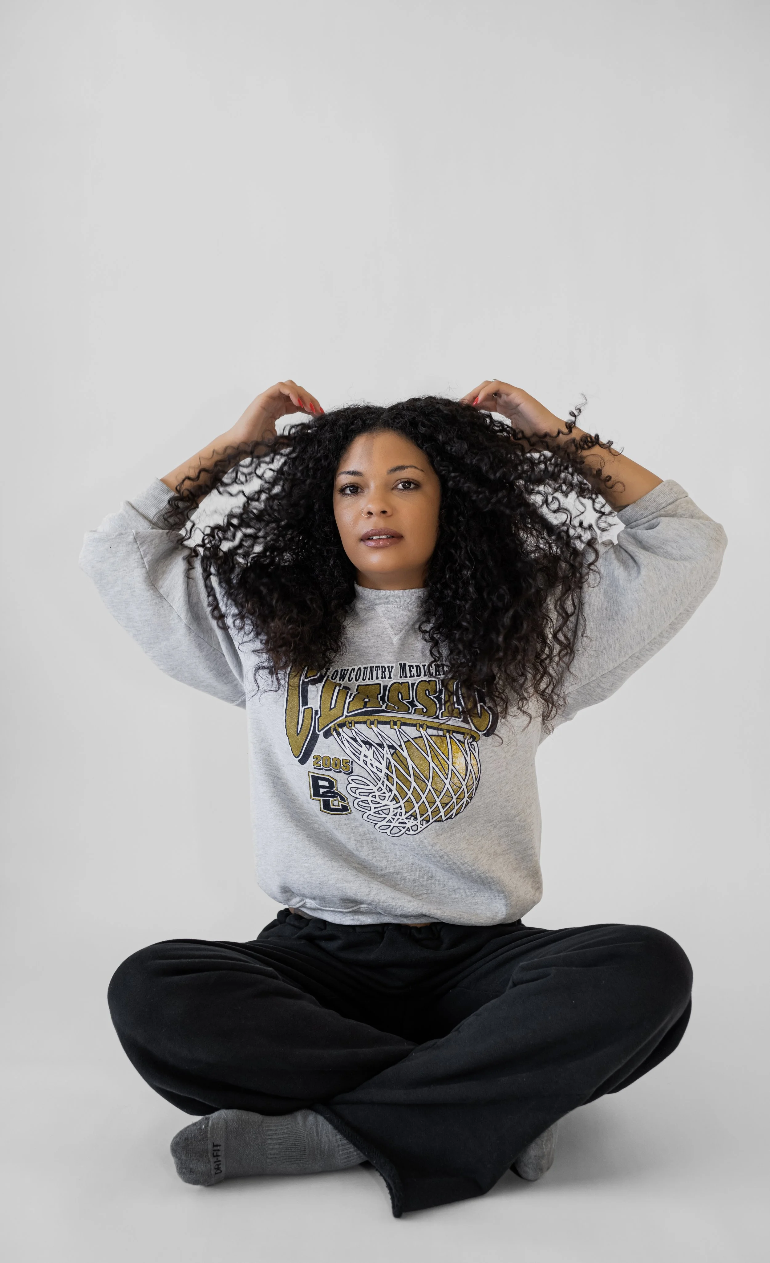 A woman with curly hair sitting cross-legged on the floor, wearing a grey sweatshirt and black pants, with her arms raised and hands behind her head.