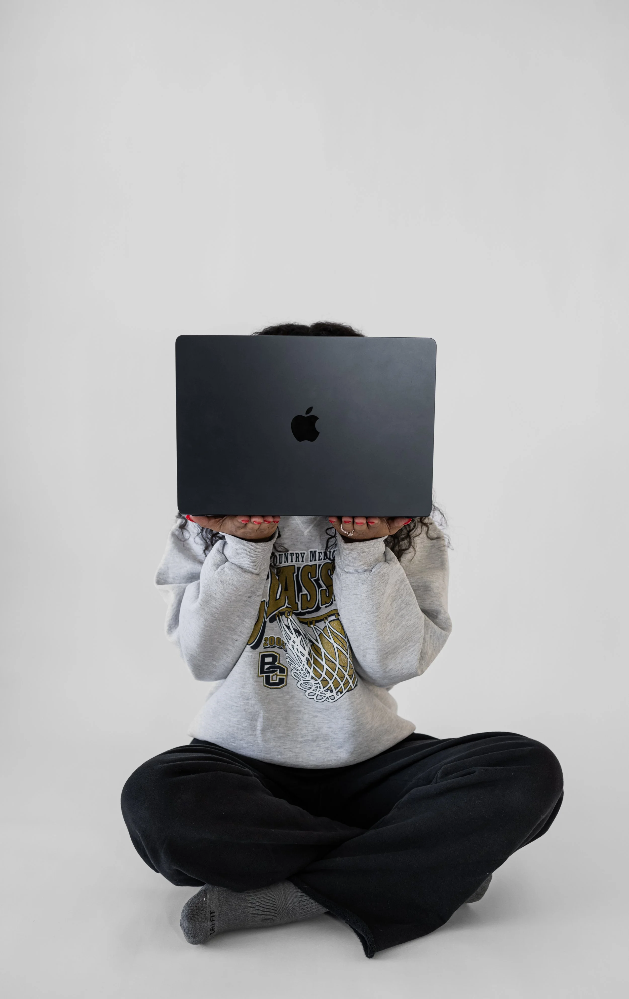 Child sitting cross-legged and holding a black laptop in front of their face, against a plain white background.