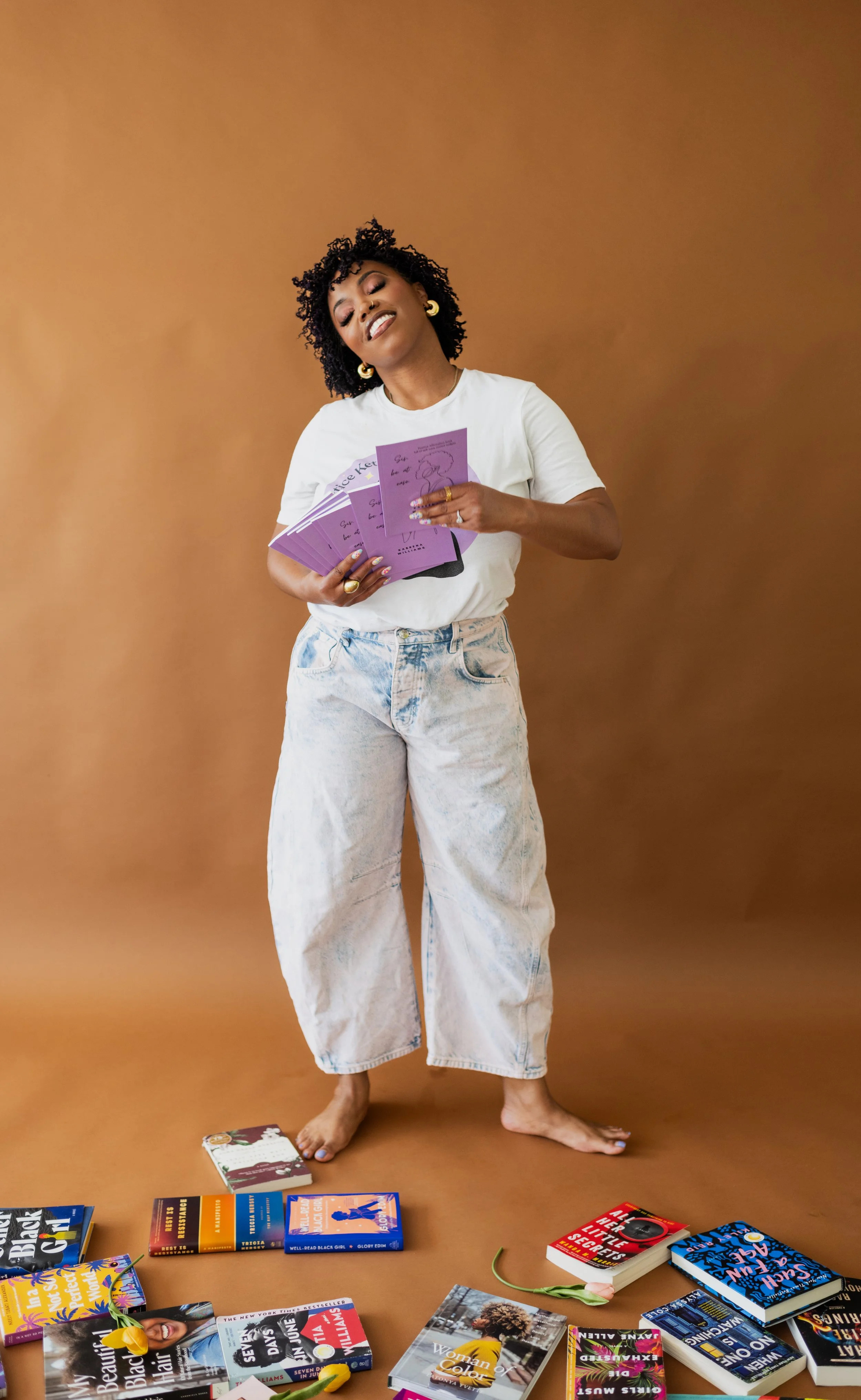 A woman standing barefoot in front of a brown background, holding several purple booklets, surrounded by various books on the floor.