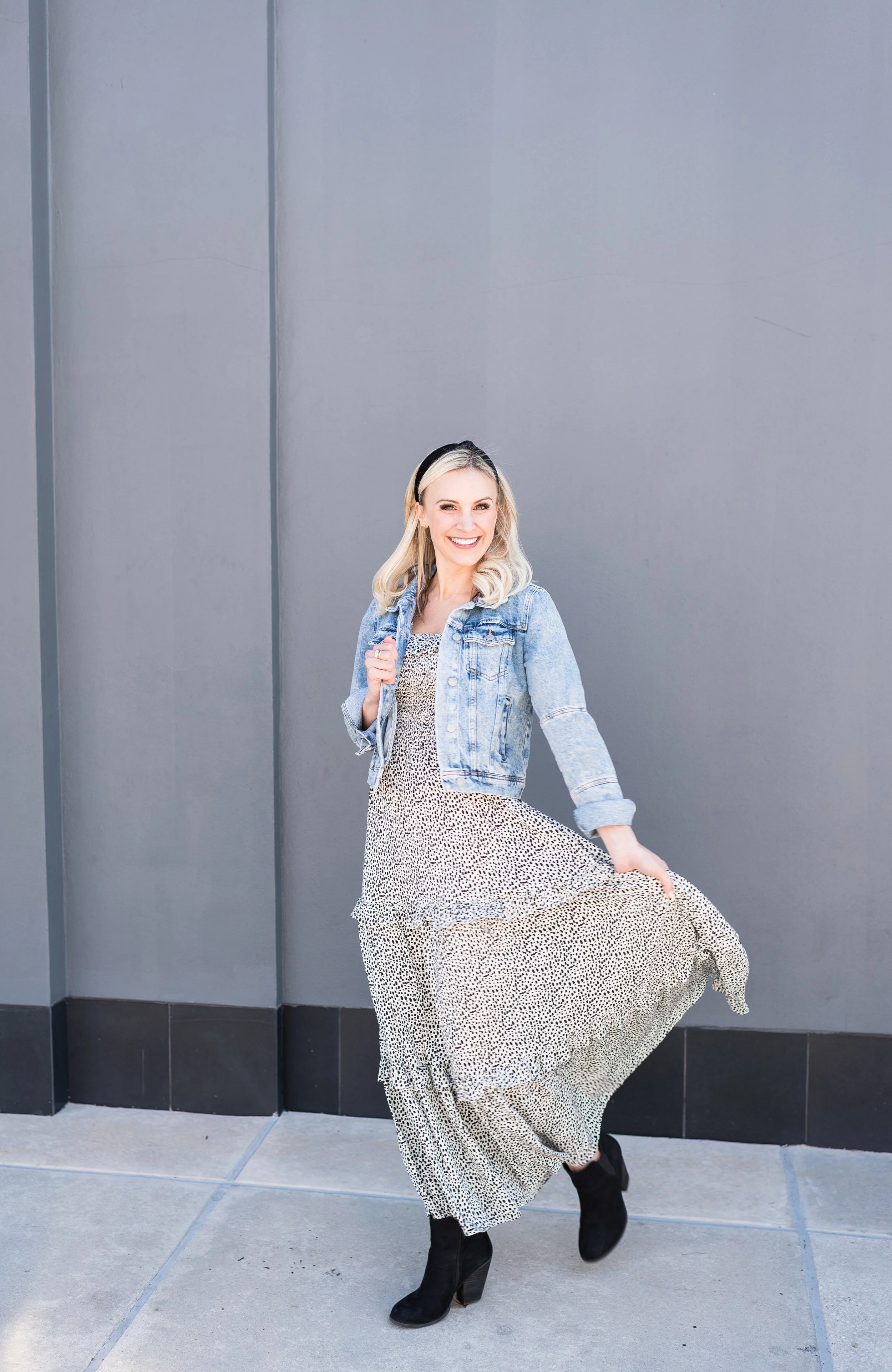 A woman with blonde hair smiling, wearing a denim jacket, a flowy animal-print dress, and black boots, standing against a gray wall.