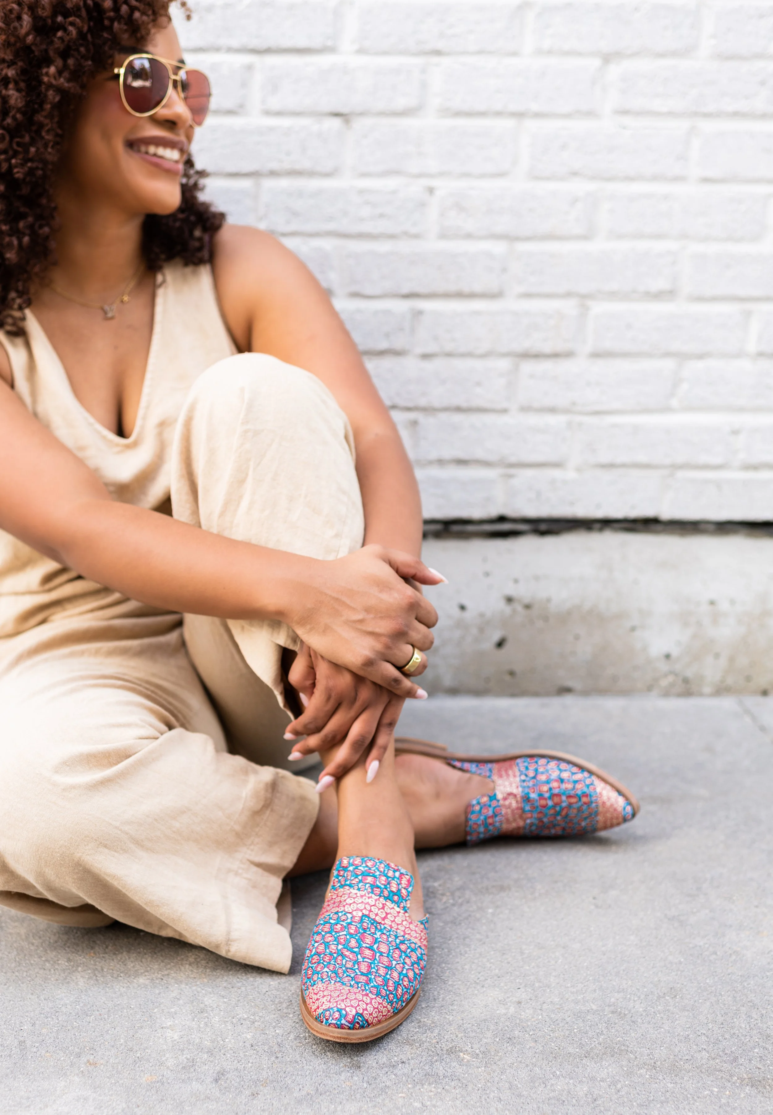 A woman with curly hair, wearing sunglasses, beige tank top, and beige pants, sitting on the ground against a white brick wall. She is smiling, with her arms wrapped around her knee, and wearing colorful patterned slip-on shoes.