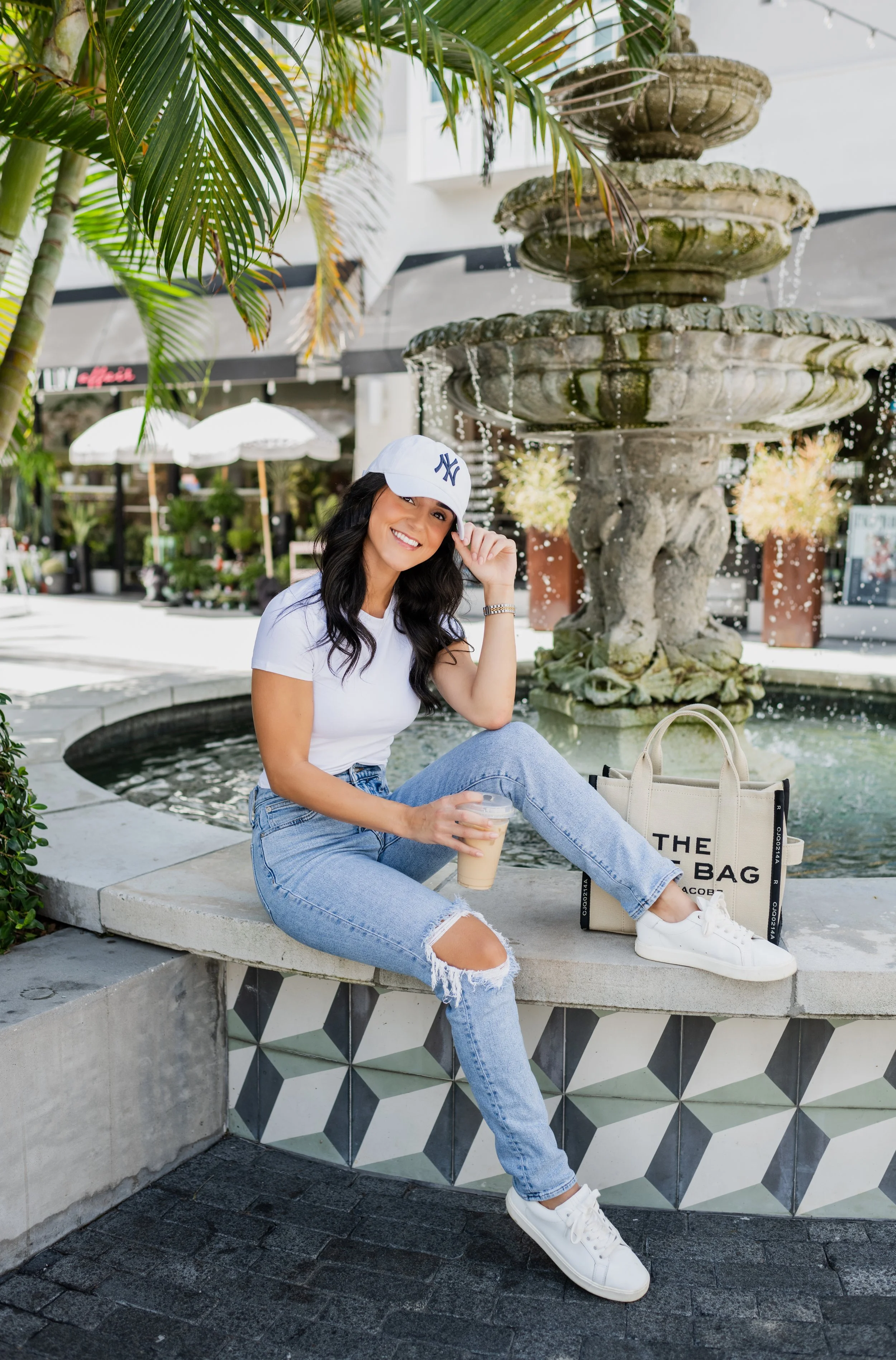 A woman sitting on a concrete bench in front of a fountain, holding a coffee drink, wearing a white cap, white t-shirt, ripped jeans, and white sneakers, with a tote bag nearby, in a shopping area with store fronts and umbrellas.