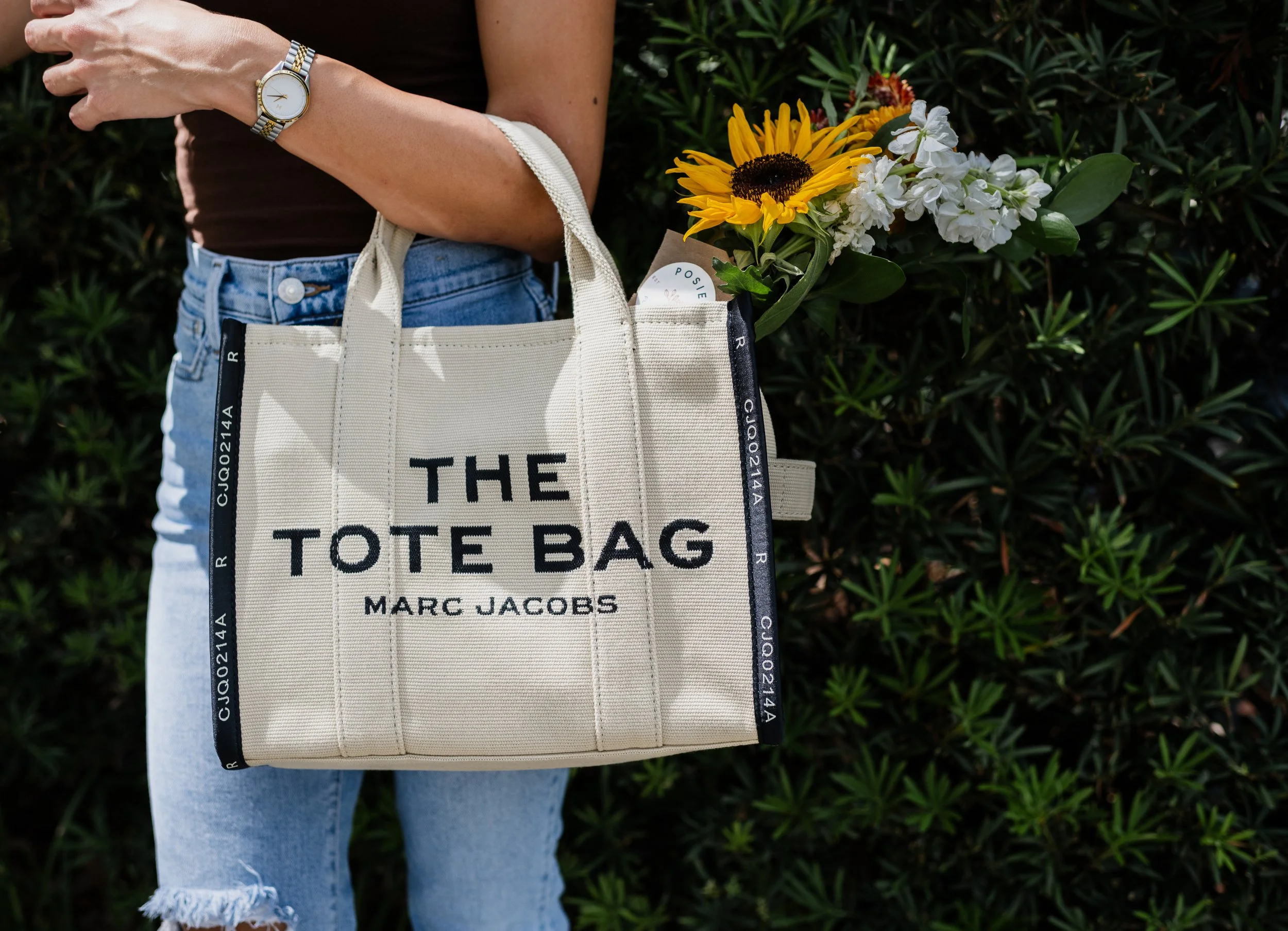 Person holding a beige Marc Jacobs tote bag with black lettering that reads 'THE TOTE BAG' and a bunch of flowers inside, standing in front of green foliage.