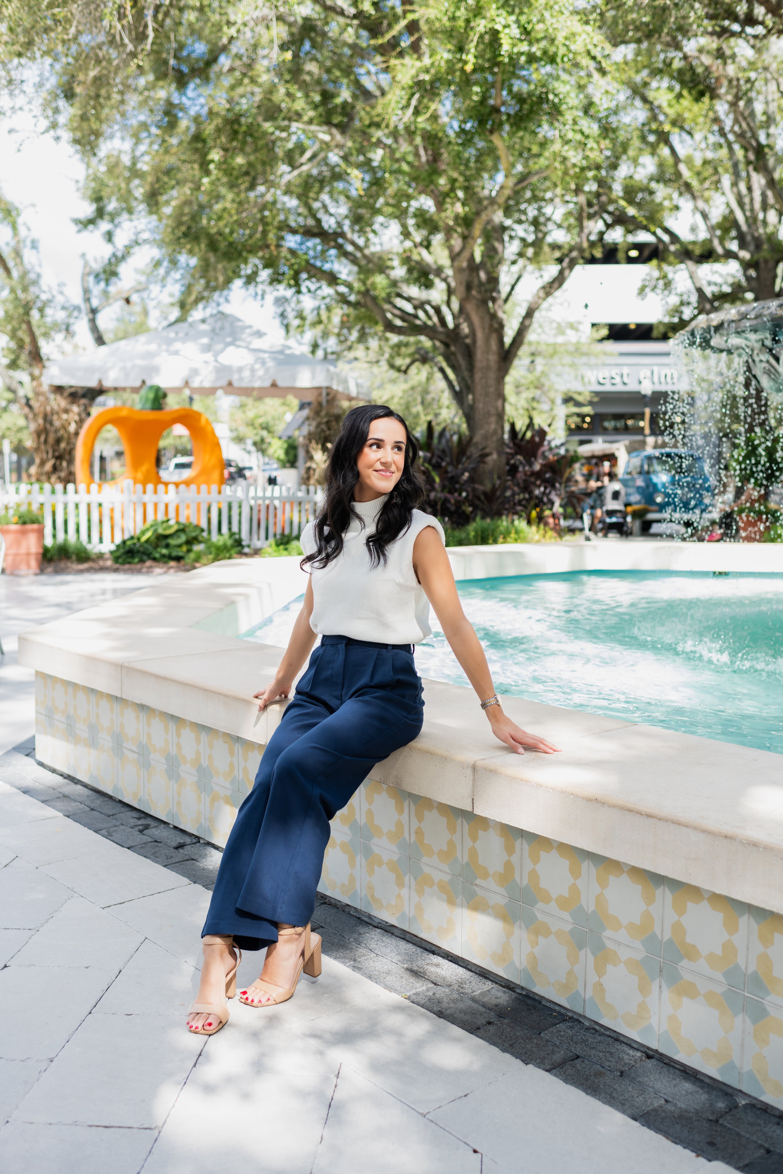 A woman sitting on a fountain's edge in an outdoor shopping area with trees, a food stand shaped like a pumpkin, and a white picket fence in the background.