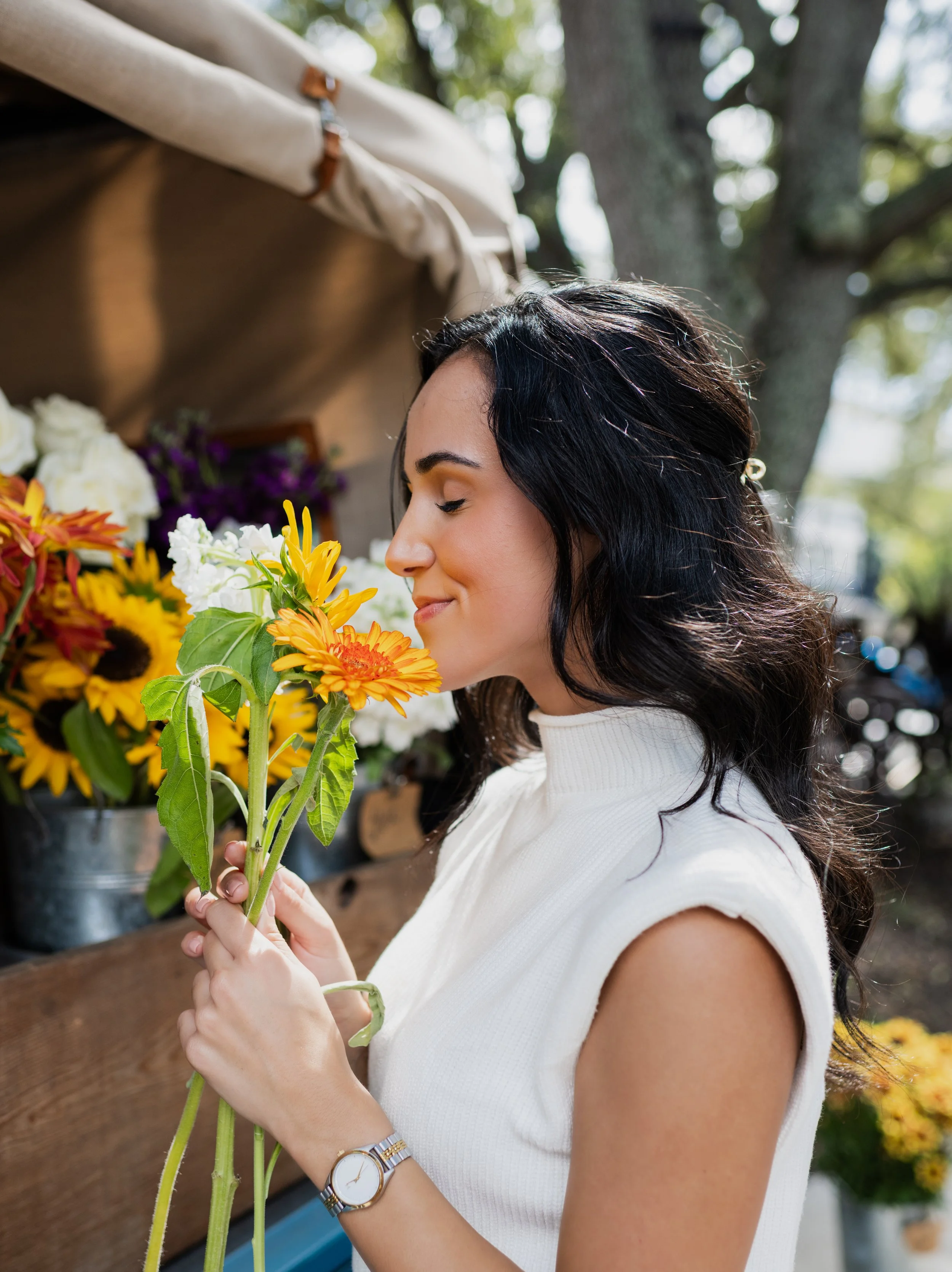 A woman selecting a bouquet of colorful flowers at an outdoor market, with trees and flowers in the background.