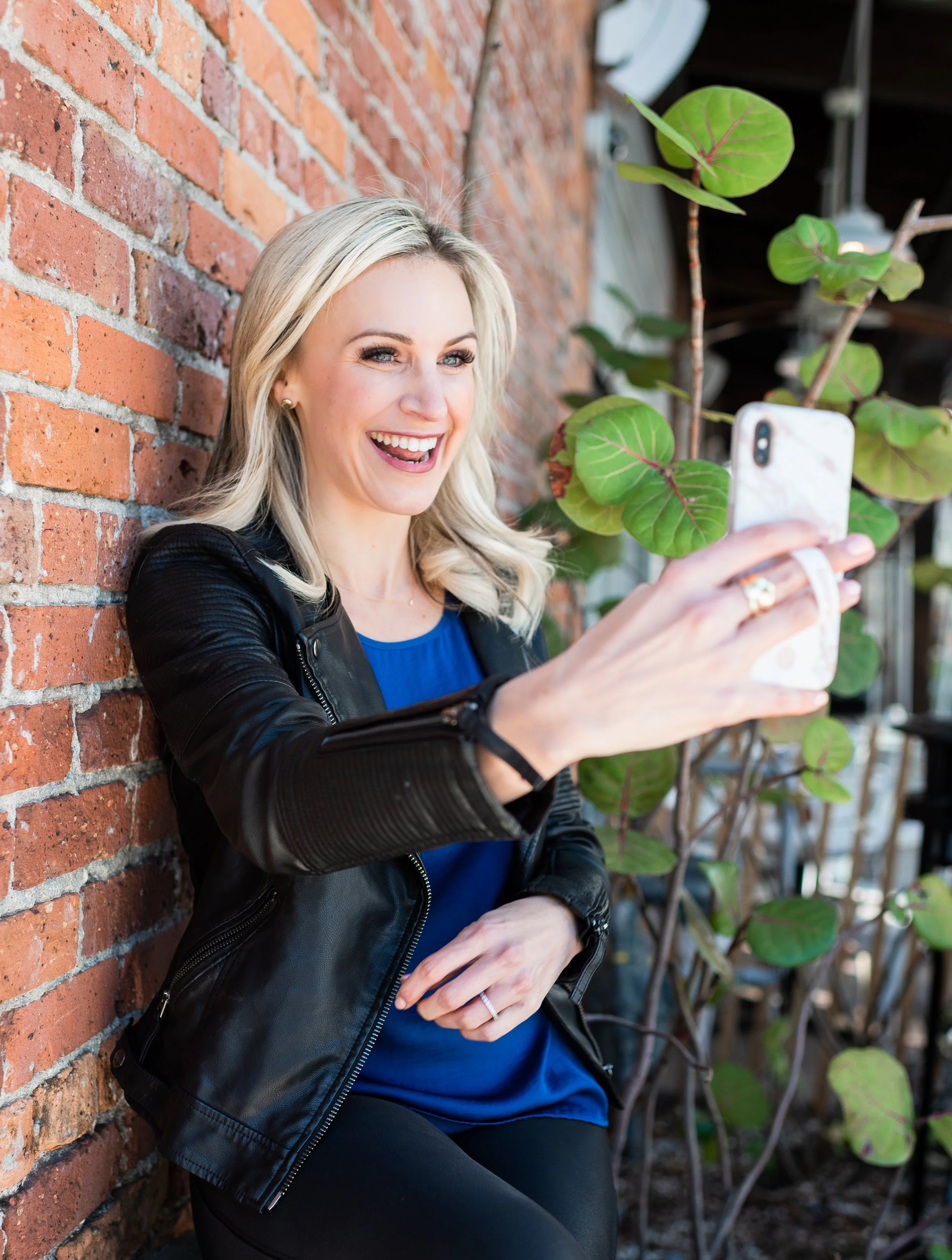 A woman with blonde hair, smiling, taking a selfie with her phone in an outdoor setting near a brick wall and green plants.