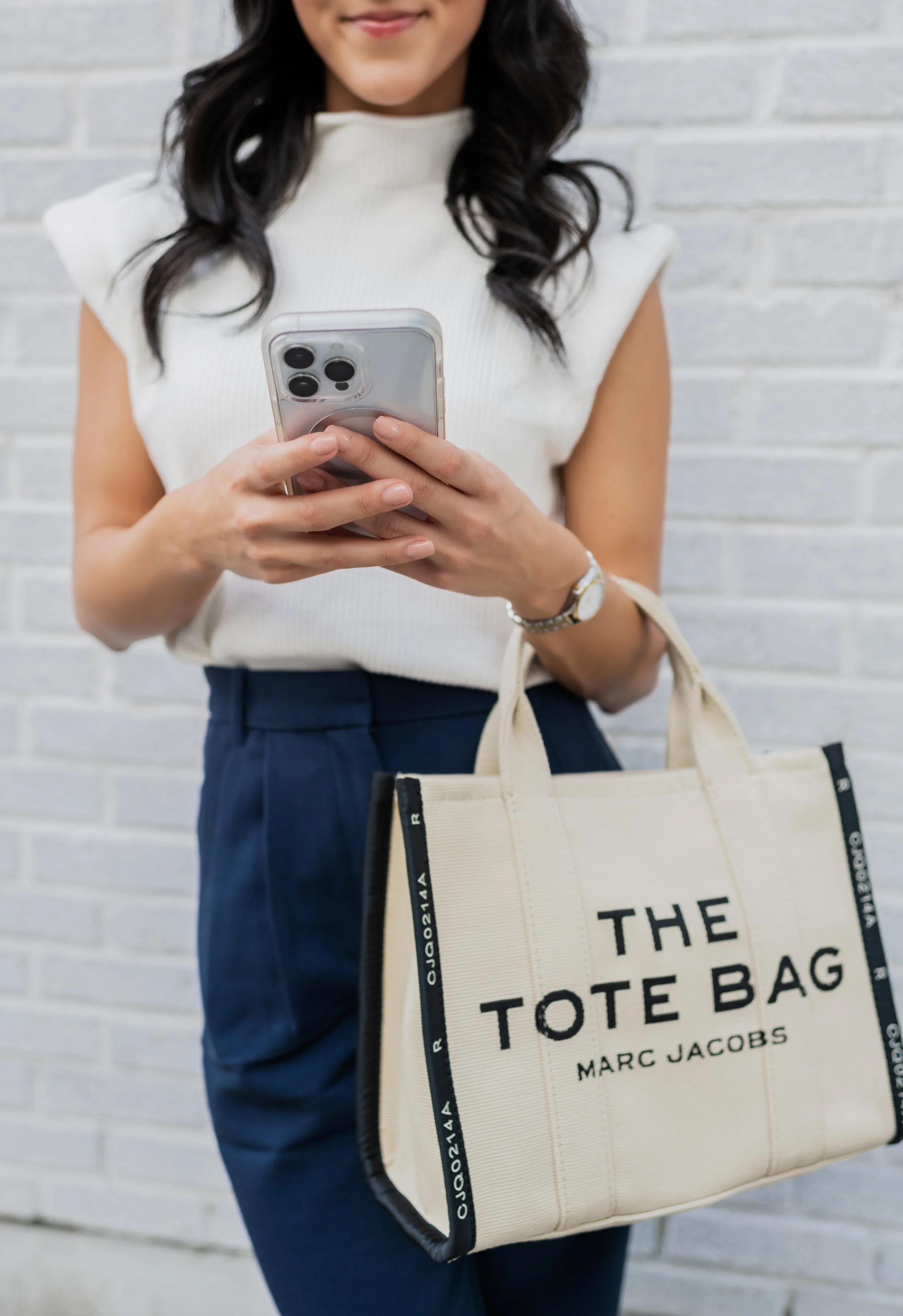 A woman holding a smartphone in front of her chest with both hands, carrying a tote bag labeled 'THE TOTE BAG MARC JACOBS'. She stands in front of a white brick wall, wearing a sleeveless cream-colored top and dark pants.