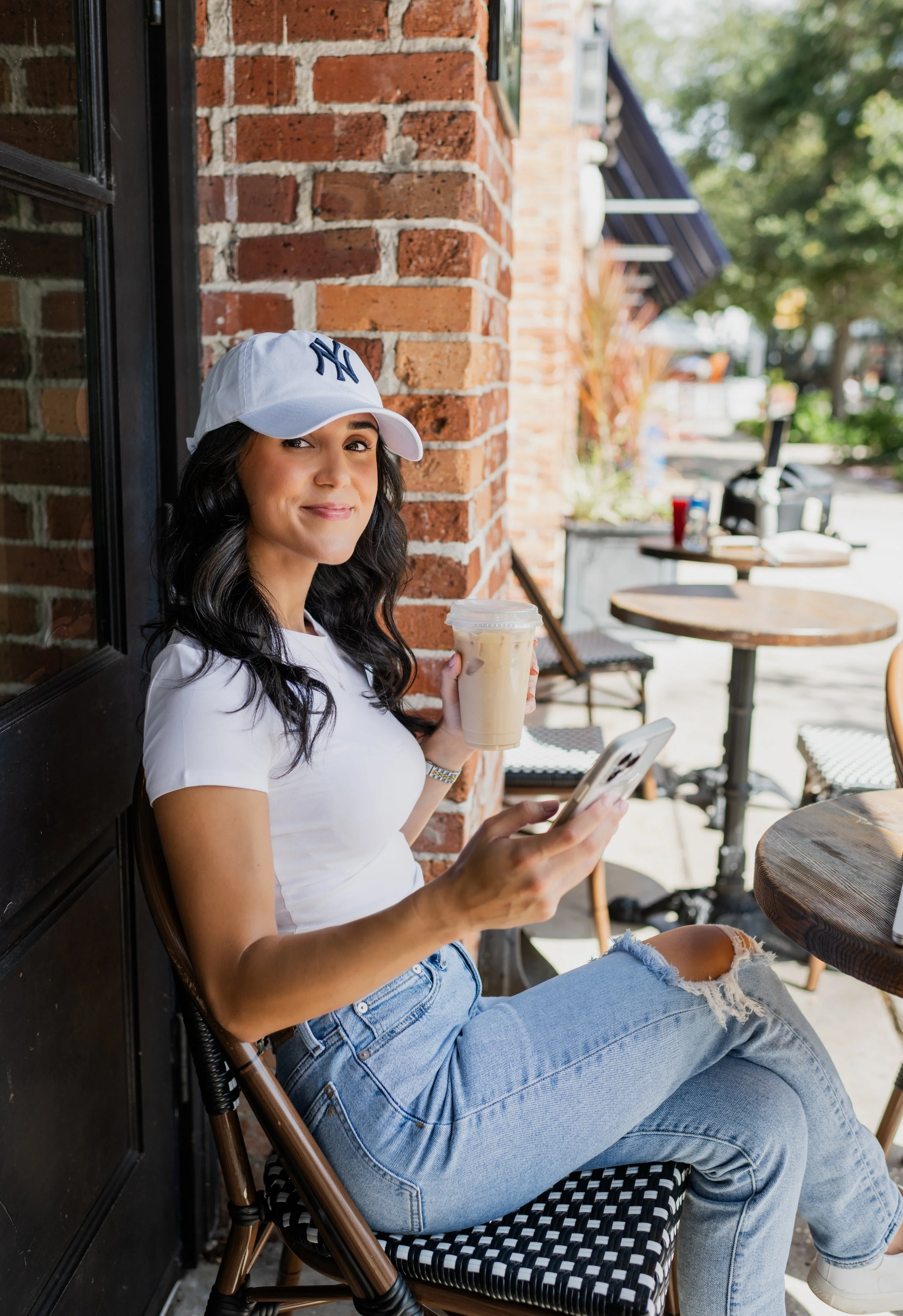 A woman sitting outside a cafe, wearing a white NY baseball cap and a white t-shirt, holding a cold coffee drink and looking at her smartphone.