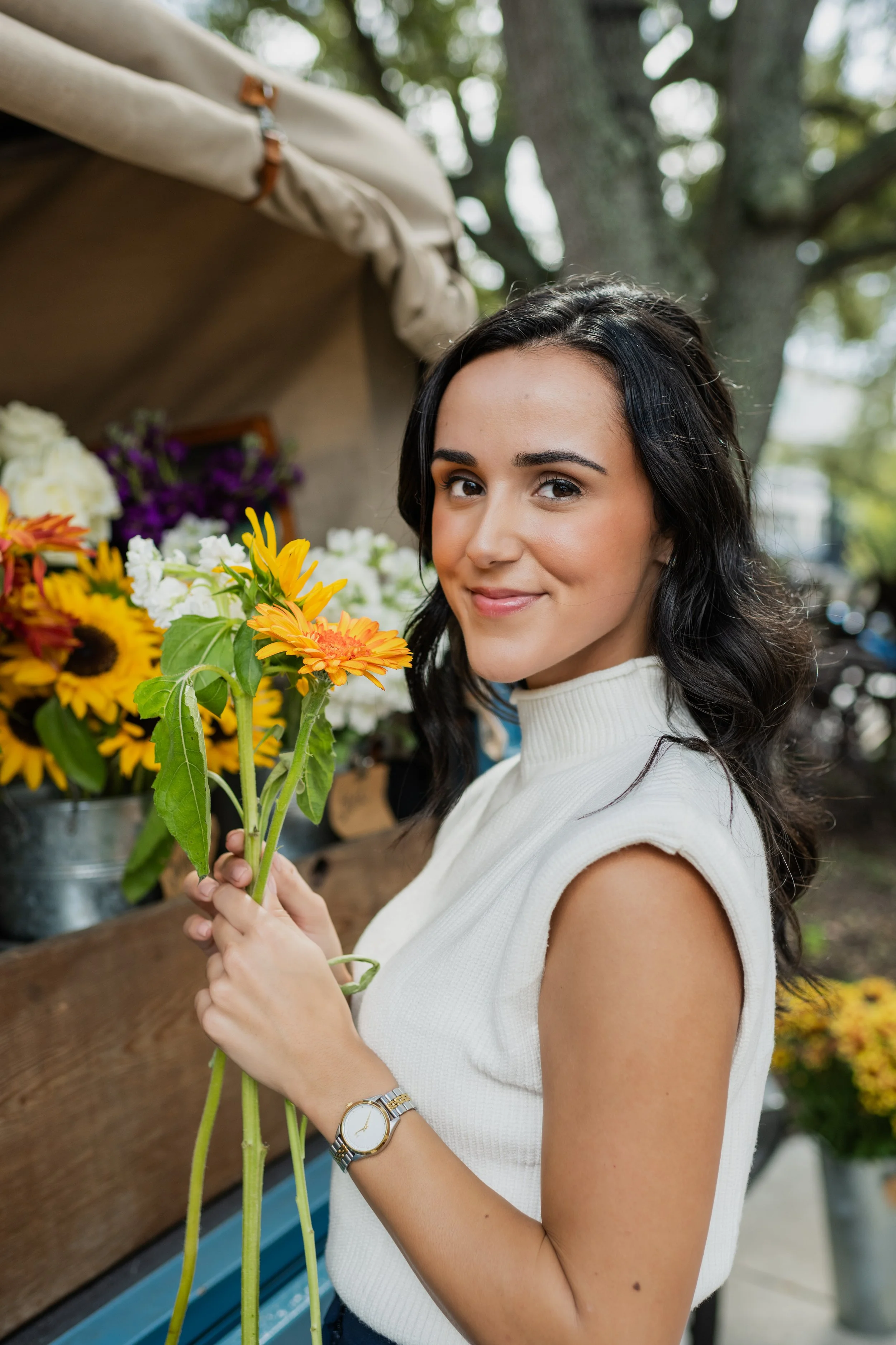 A woman with dark hair holding a bunch of yellow and orange flowers, standing near a flower stall outdoors.