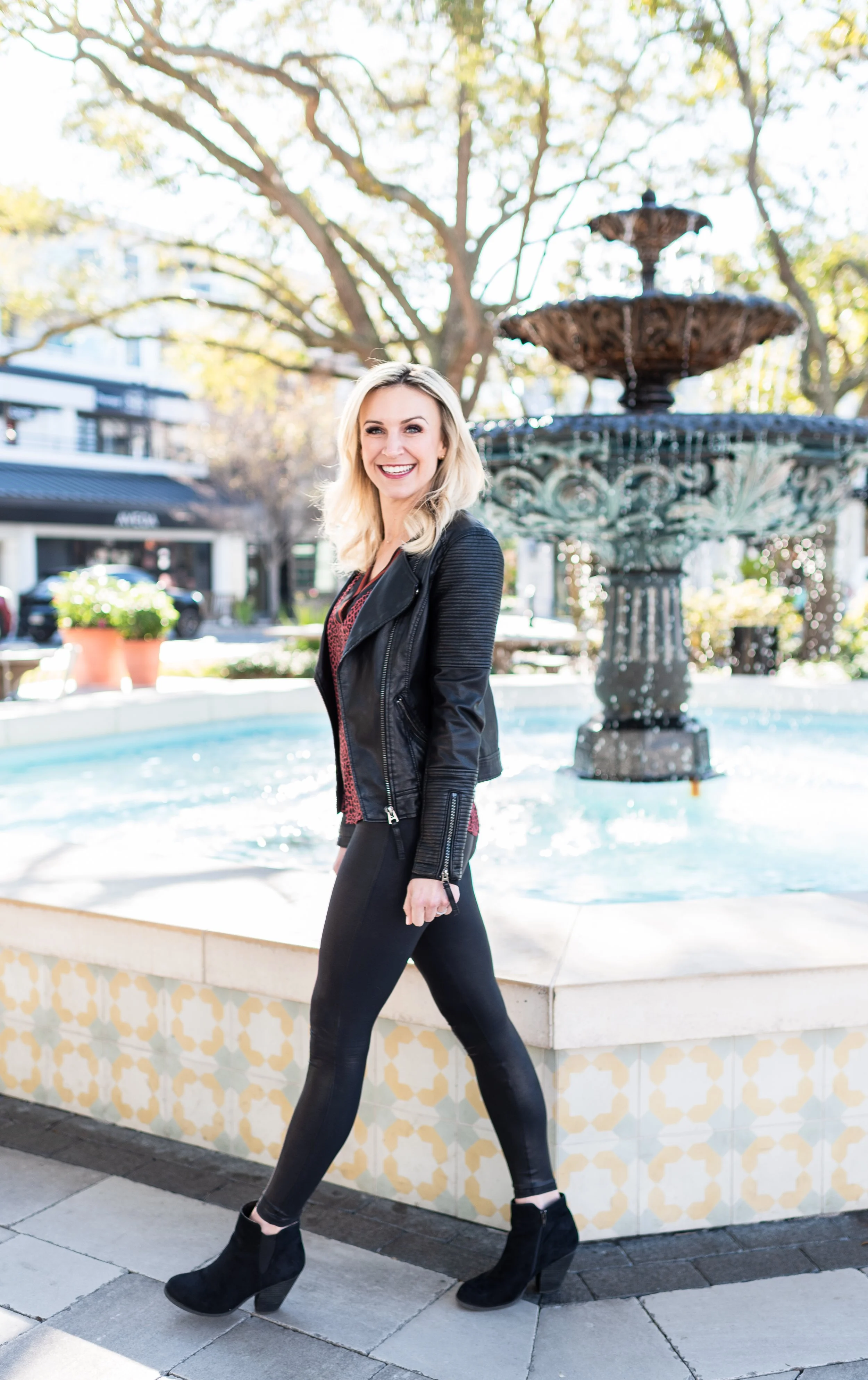 A woman with blonde hair smiling and walking past a fountain in an outdoor urban setting, wearing a black jacket, red top, black leggings, and black ankle boots.