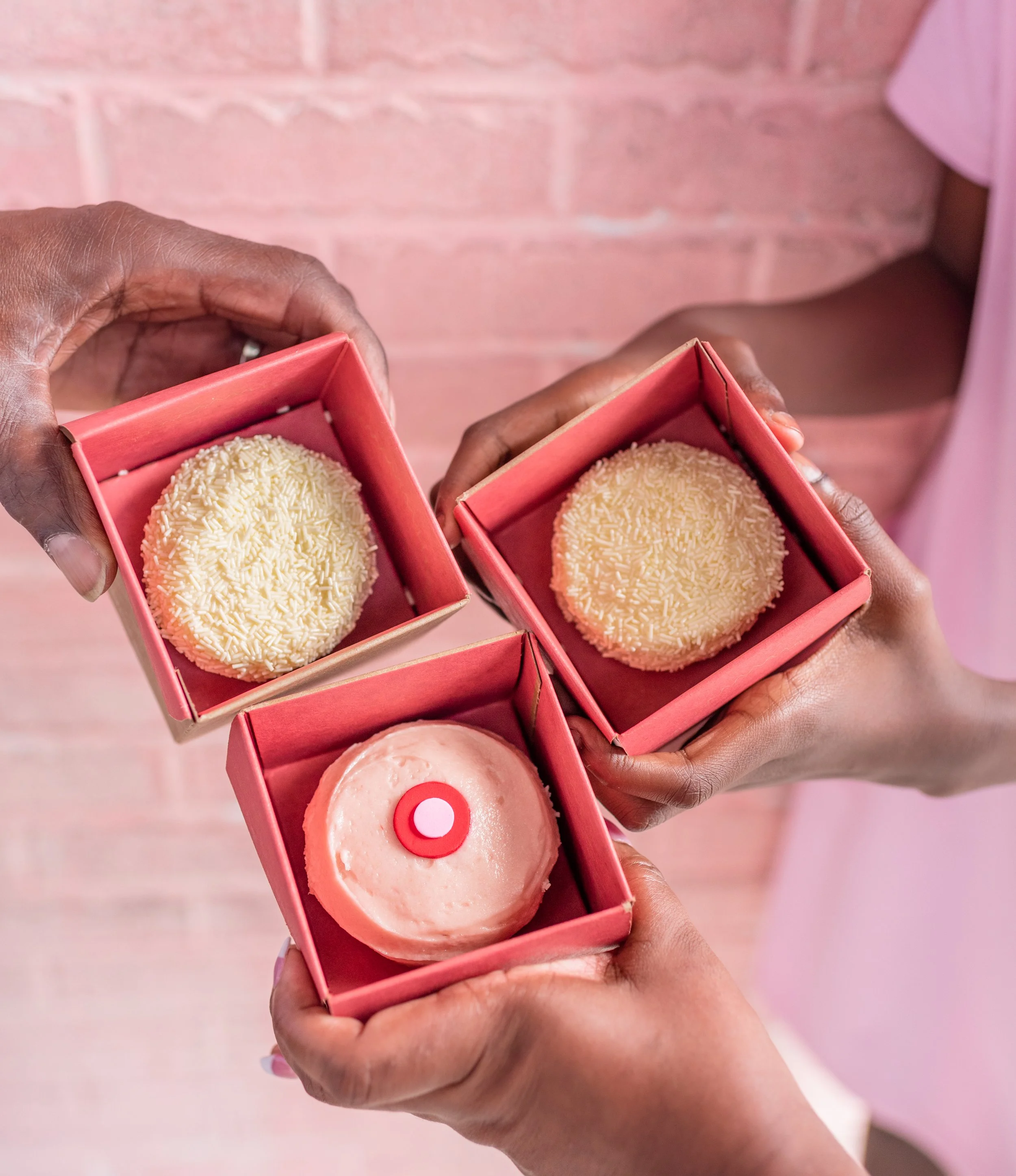 Three hands holding small pink boxes, each containing a round dessert. Two of the desserts are covered in white sprinkles, and one has a pink frosted icing with a red and white edible decoration on top.