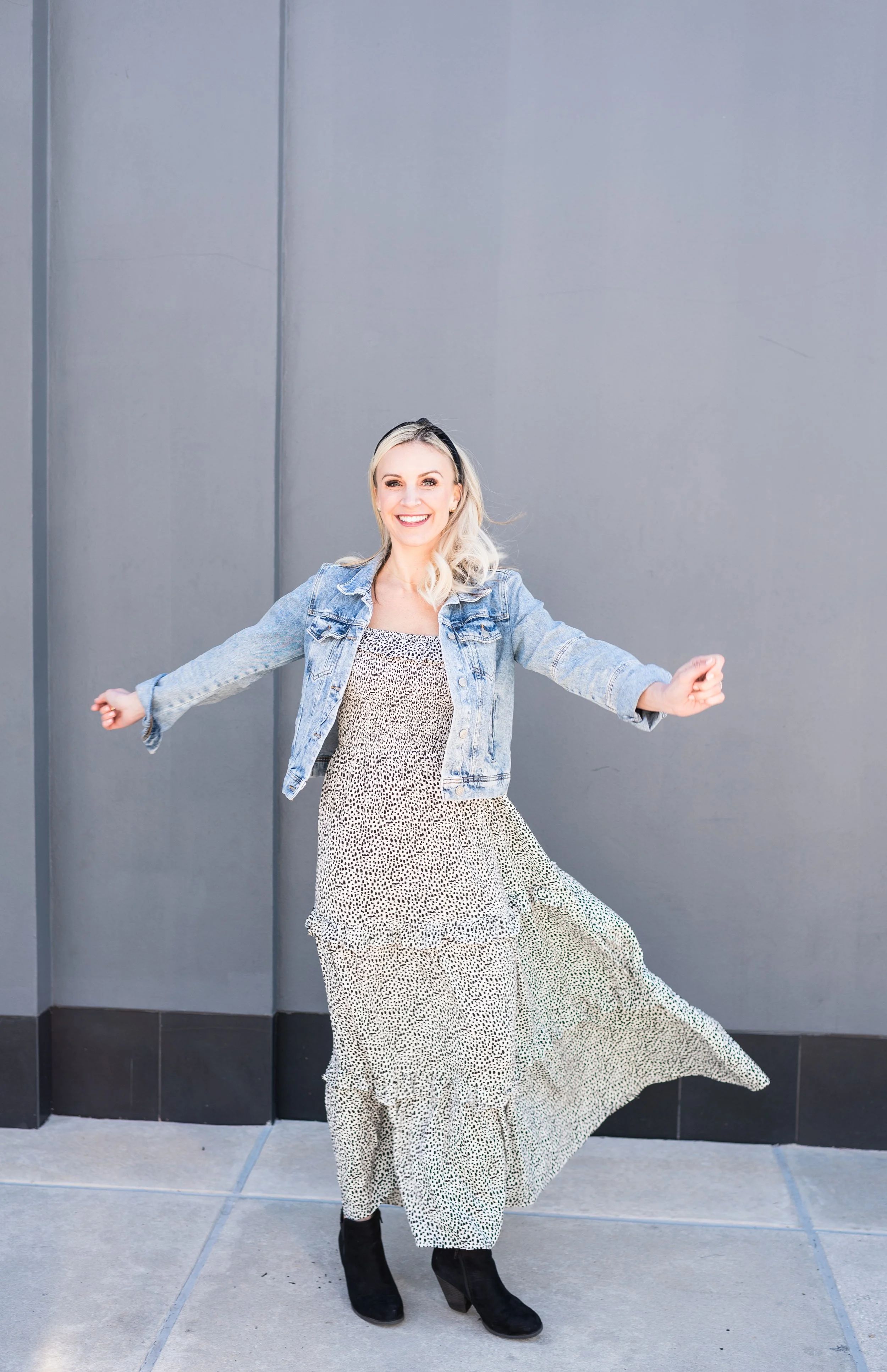 A woman with blonde hair wearing a denim jacket, a patterned maxi dress, and black boots, smiling and twirling outdoors against a grey wall.
