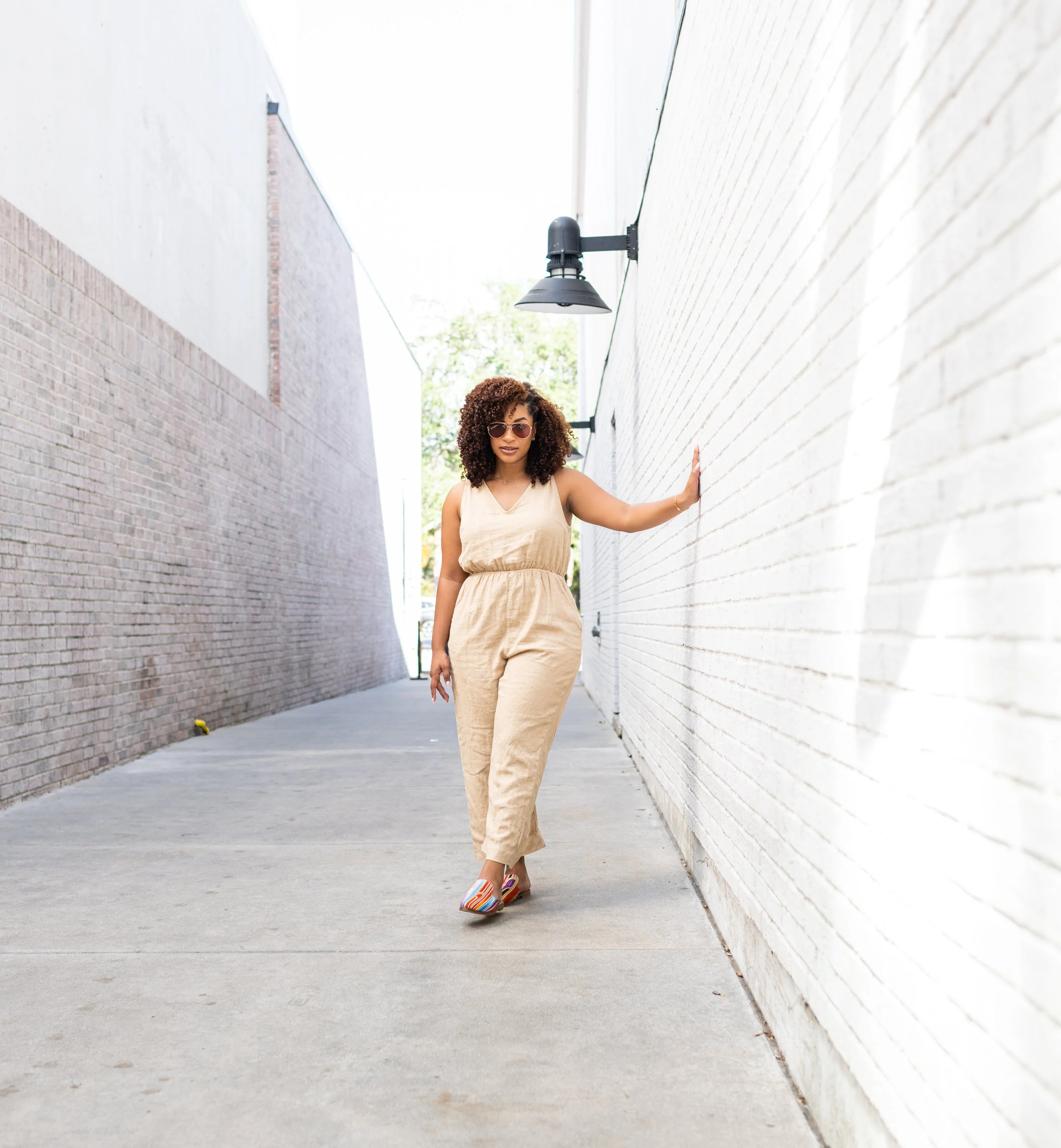 A woman with curly hair and sunglasses in a beige jumpsuit walking in an alleyway, touching a white brick wall on her right.