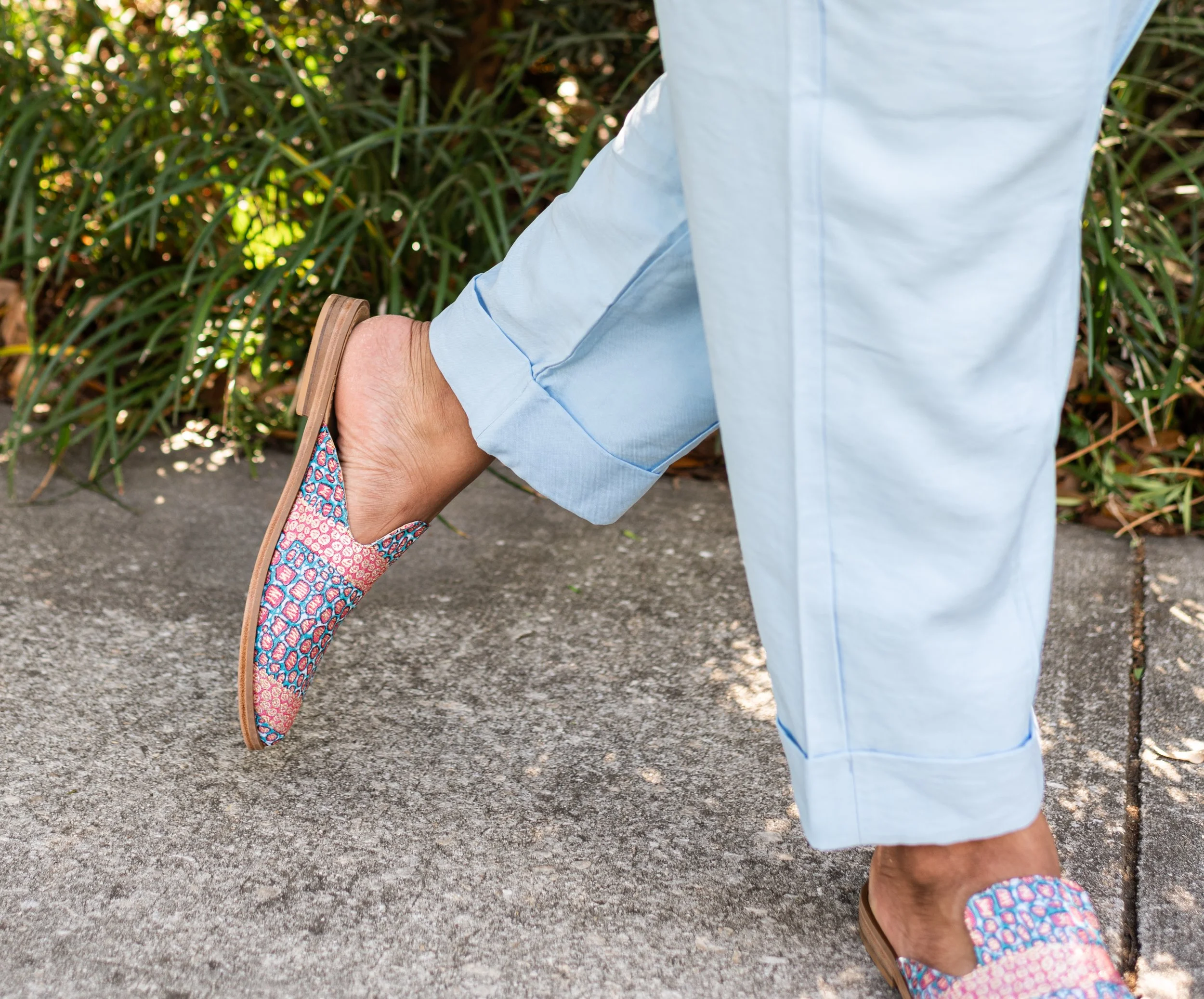 Person walking outdoors wearing light blue pants and colorful patterned slip-on shoes