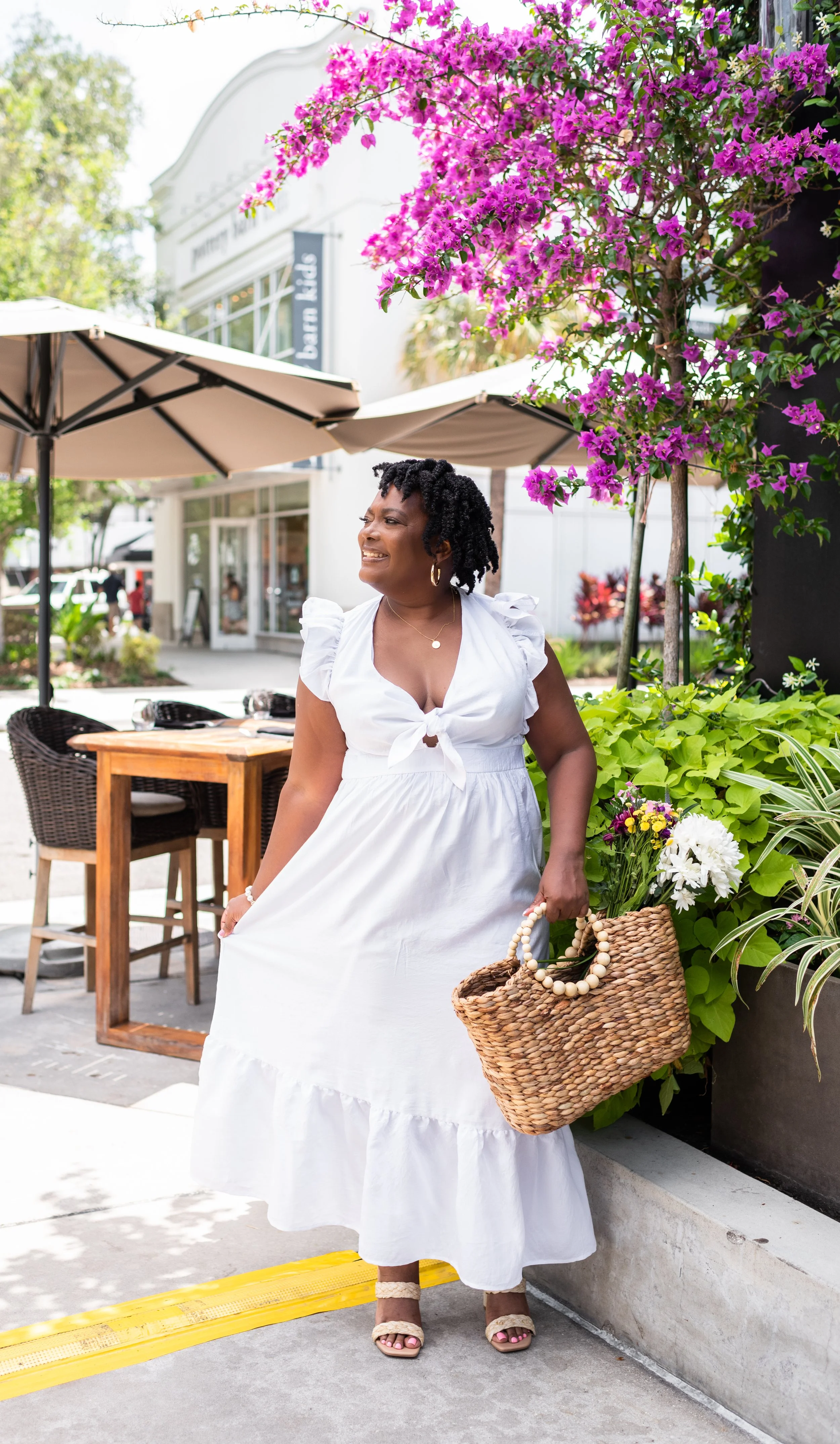 A woman in a white summer dress holding a wicker basket with flowers, standing outdoors in front of a lush plant and purple flowers, with outdoor furniture and a building in the background.