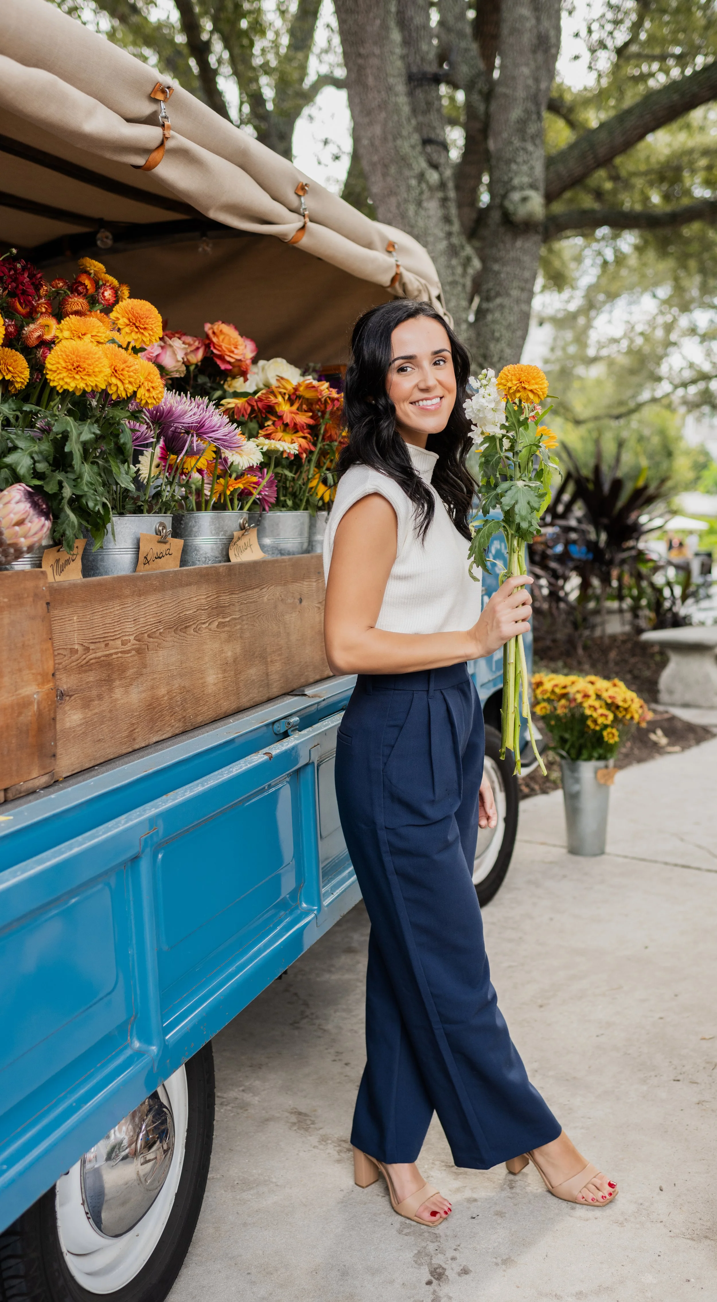Woman smiling and holding yellow and white flowers next to a flower stand with colorful blooms, in an outdoor setting with trees.
