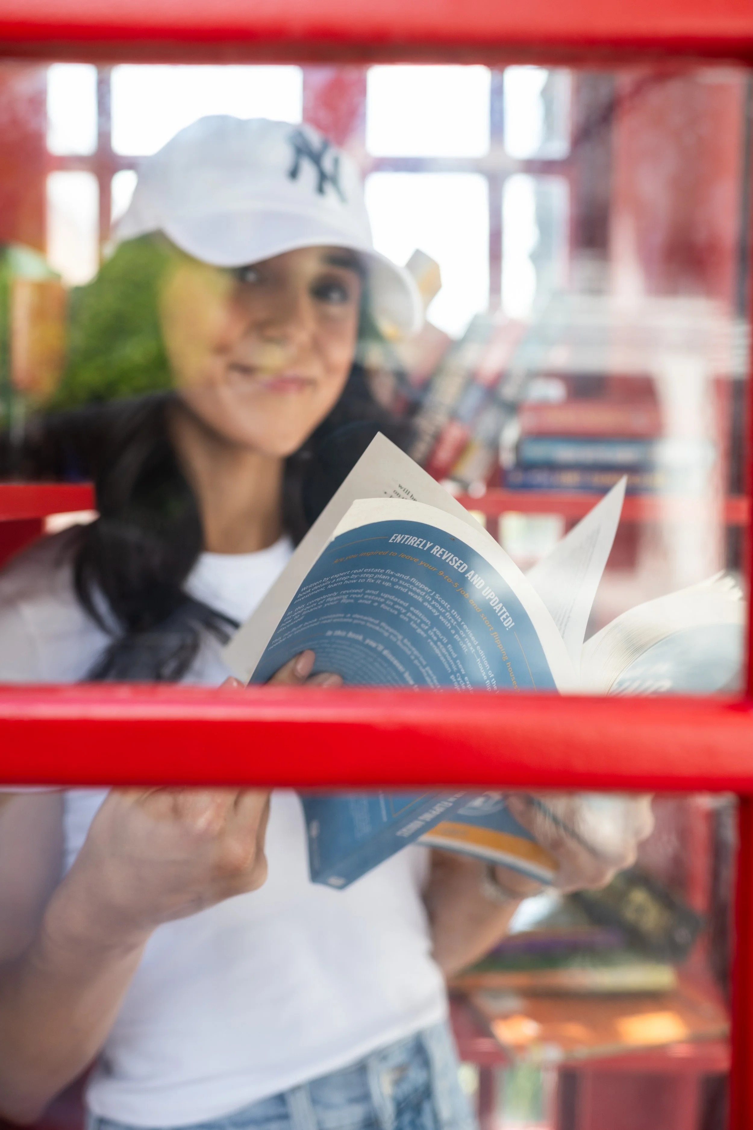 Girl wearing a white cap and white t-shirt, looking through a red window frame while reading a blue informational brochure inside a bookstore or library.