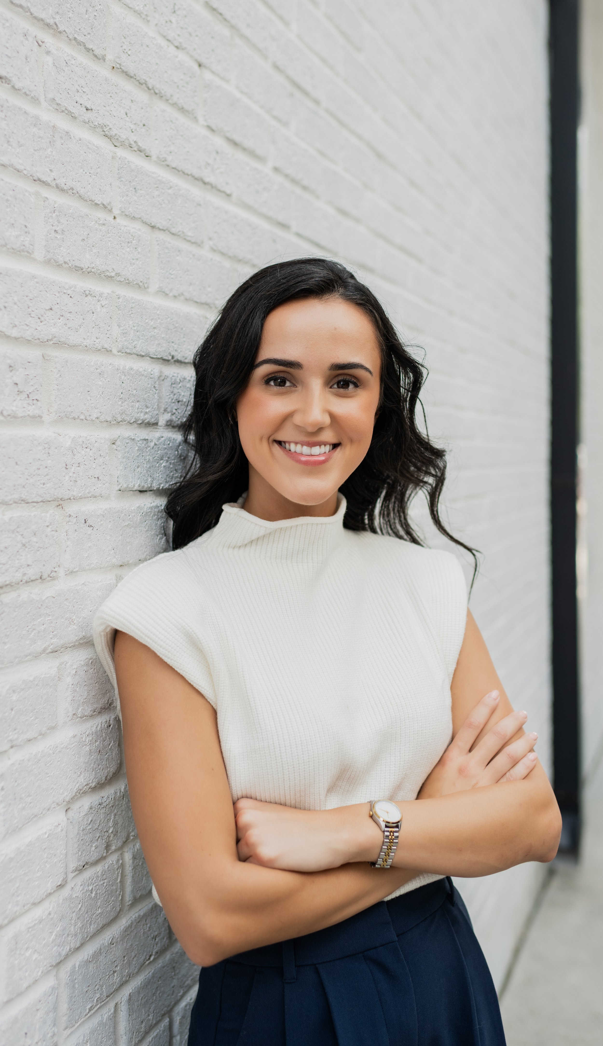 A smiling woman with dark hair and a white sleeveless top standing against a white brick wall with her arms crossed.
