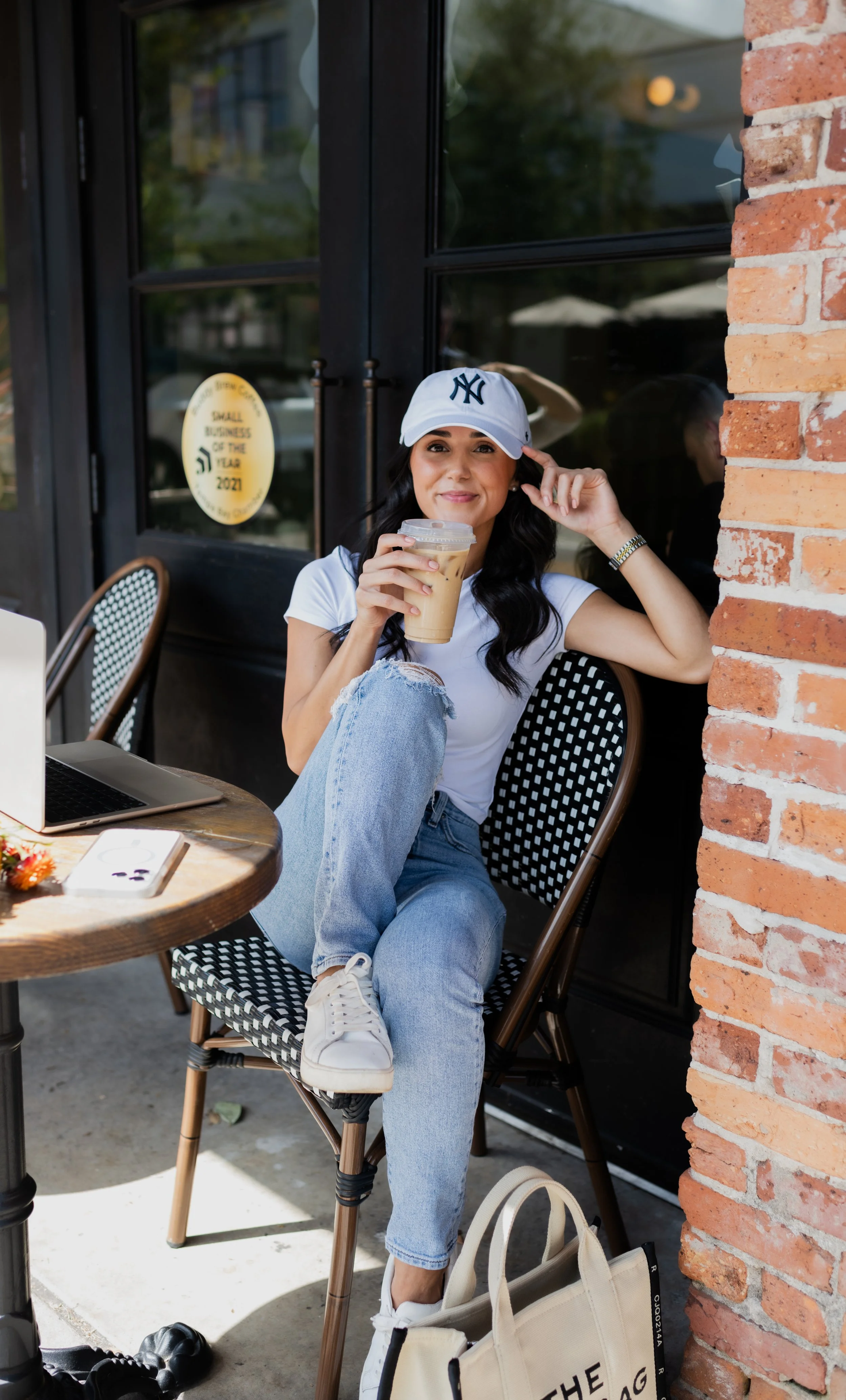 A woman sitting outside a cafe, wearing a white baseball cap, holding a coffee drink, and smiling at the camera.