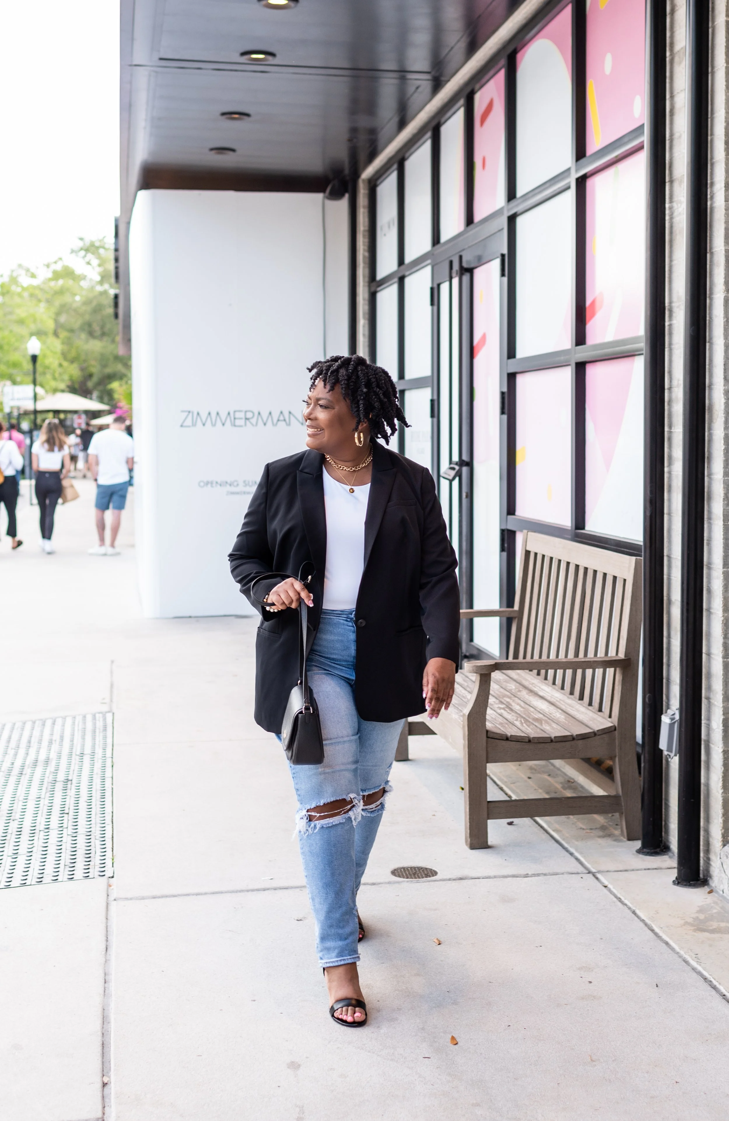 Woman walking outdoors on sidewalk near a bench, wearing a black blazer, white top, ripped jeans, and black sandals, with a black handbag, smiling, with stores and people in the background.