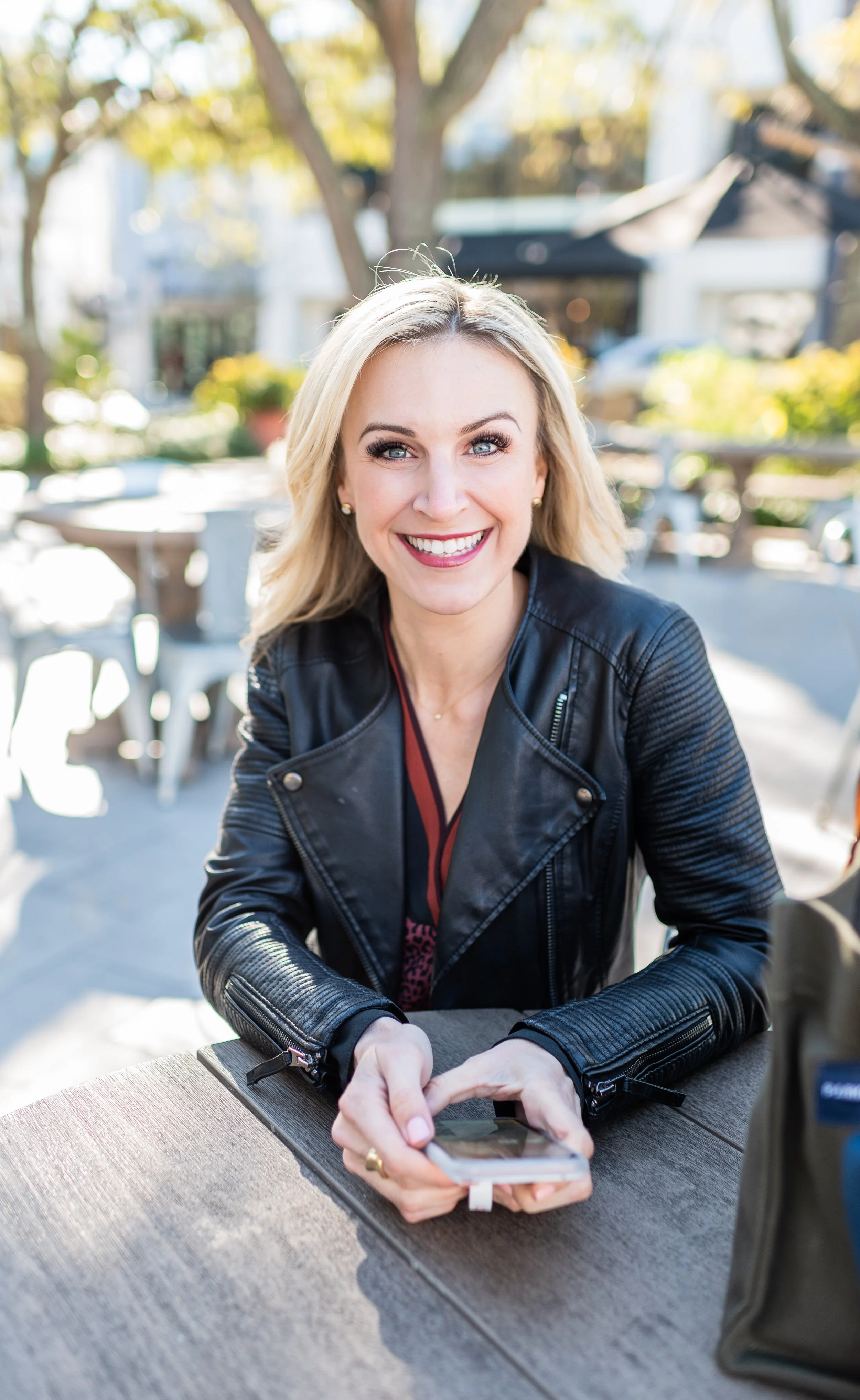 A woman with blonde hair, wearing a black leather jacket, smiling while looking at the camera and holding a smartphone at an outdoor cafe on a sunny day.