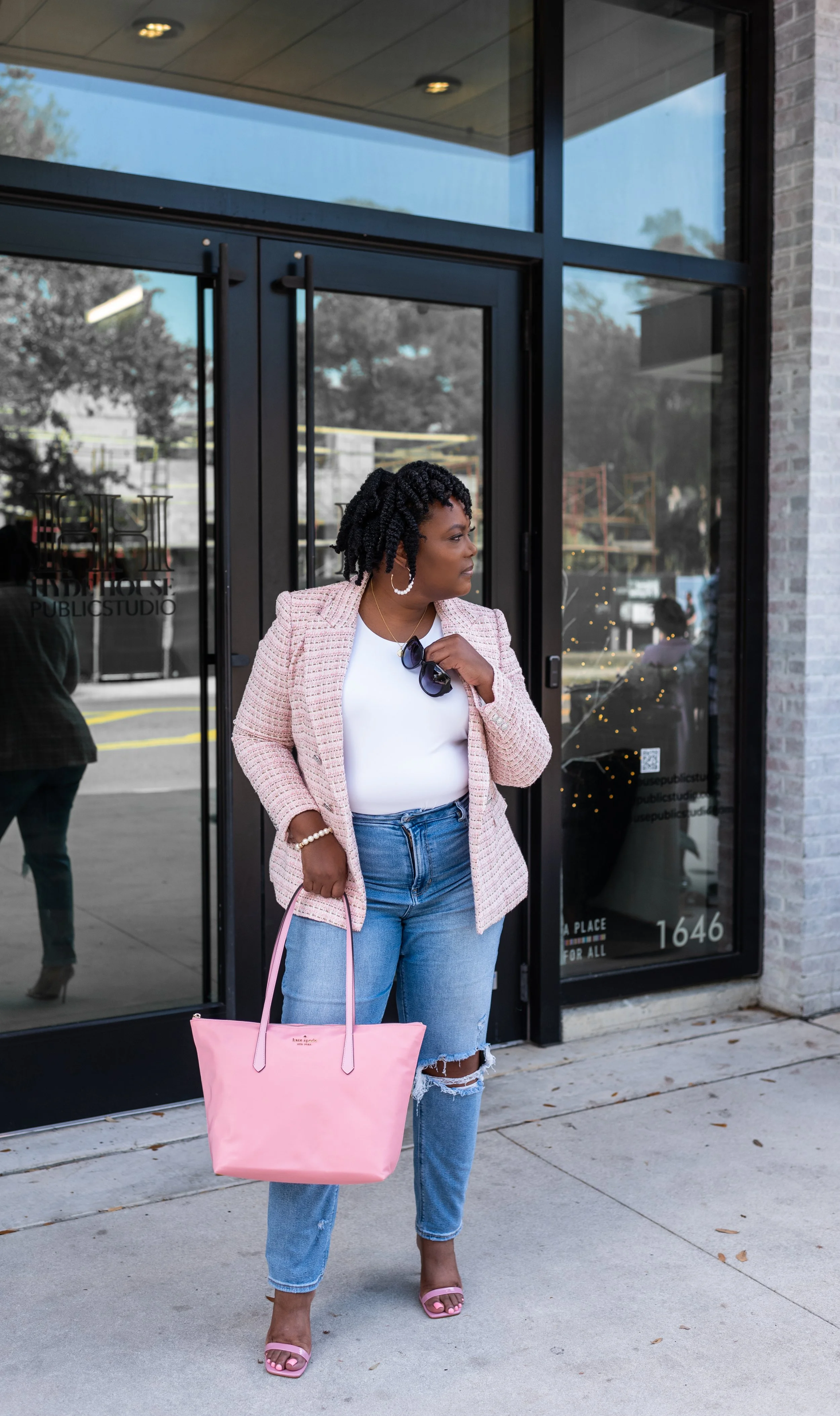 A woman standing outside a building with glass doors, holding a pink tote bag, wearing a pink blazer, white top, ripped jeans, and pink heels, with sunglasses hanging from her shirt collar.