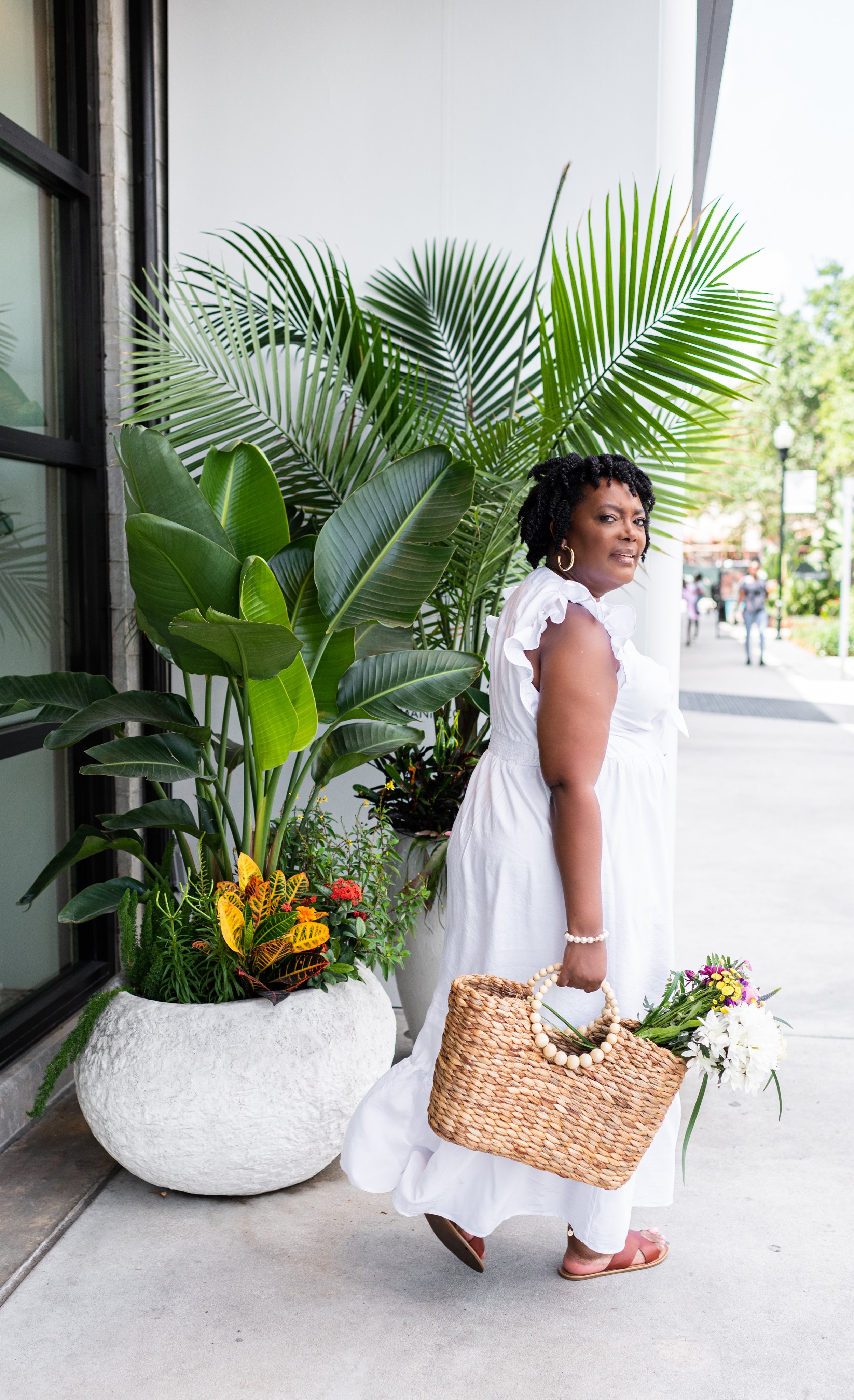 A woman in a white dress holding a woven basket with flowers, standing beside large green tropical plants outdoors.