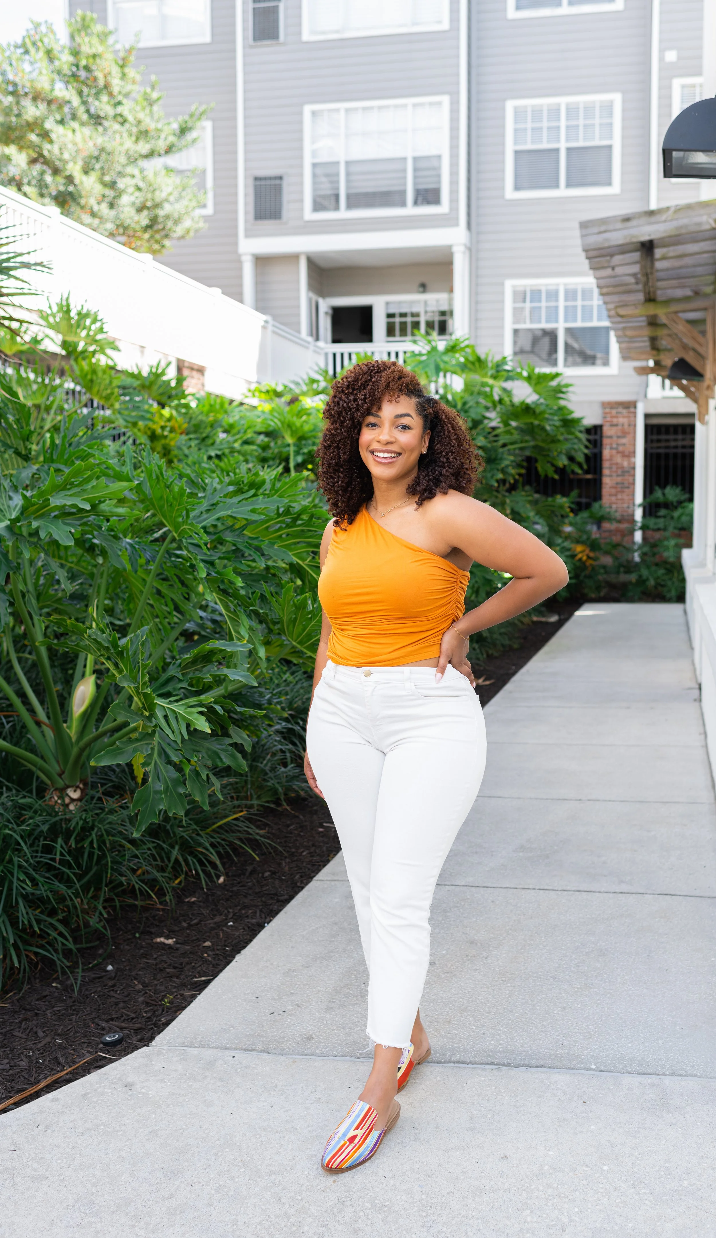 A woman in an orange top and white pants standing on a sidewalk, smiling, with green plants and a residential building in the background.