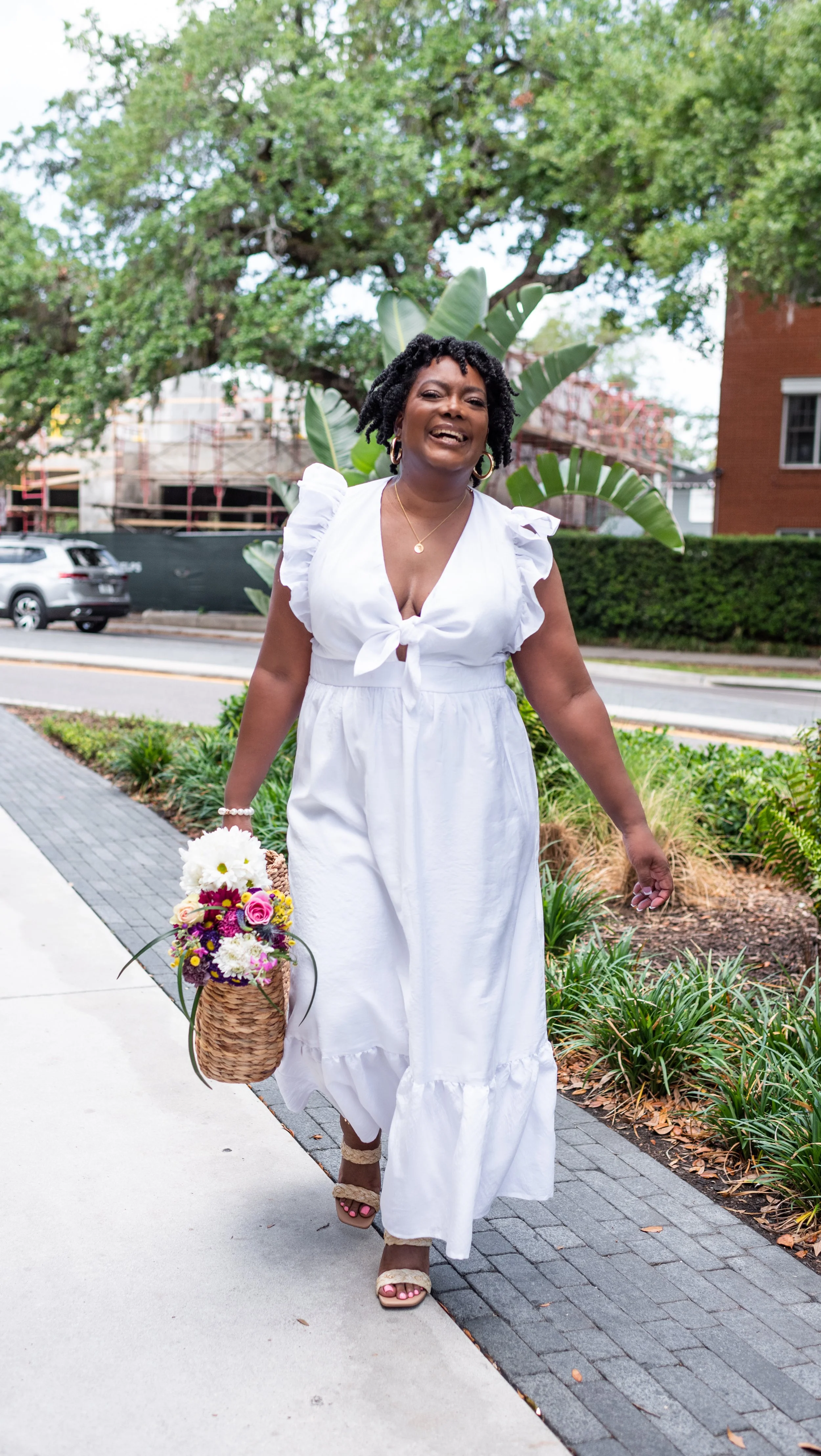 A woman in a white dress walking on a sidewalk, holding a basket of colorful flowers, with greenery and trees in the background.