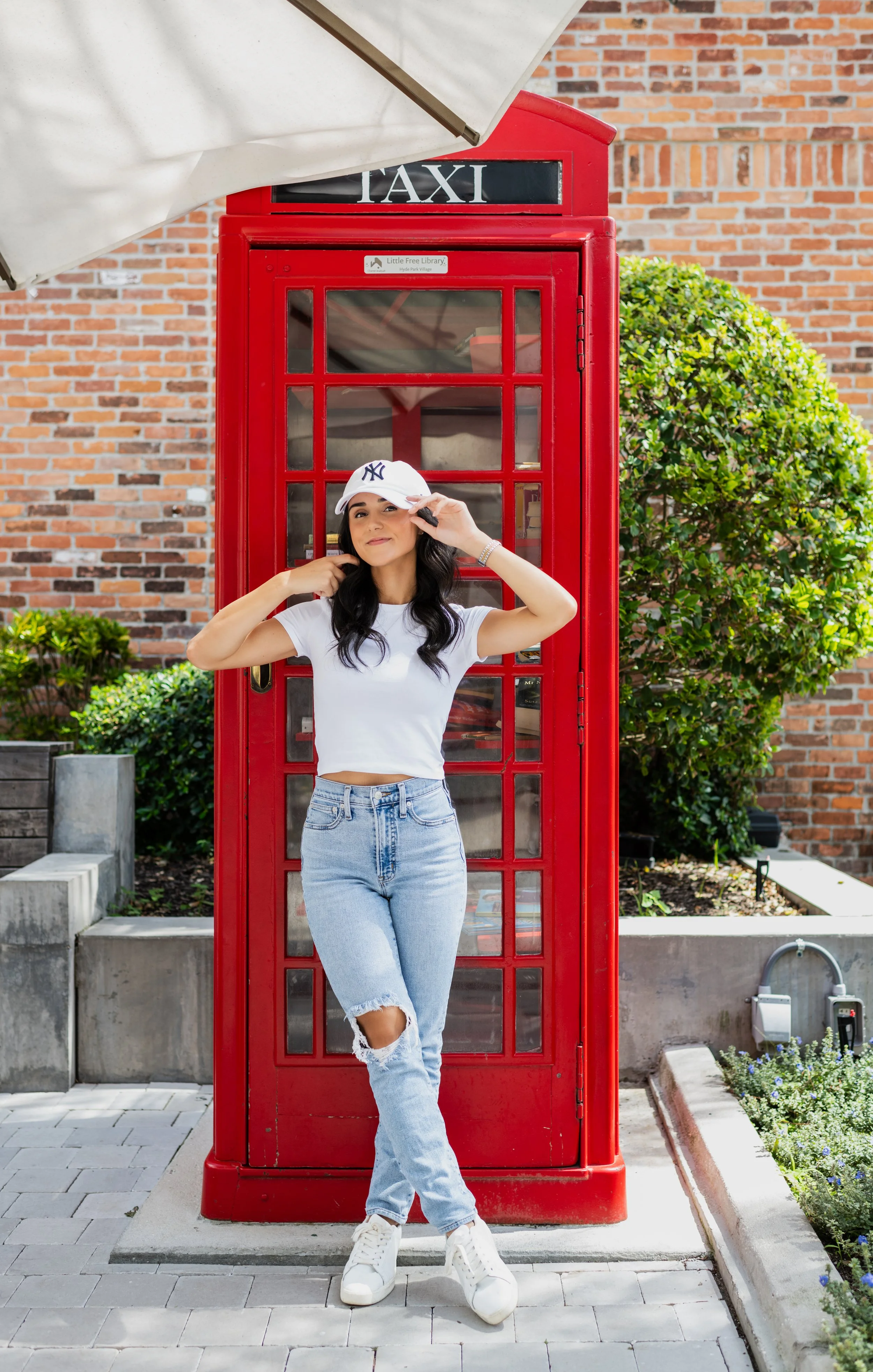 A young woman in a casual outfit, wearing a white crop top, ripped jeans, white sneakers, and a baseball cap, standing in front of a classic red telephone booth. She is adjusting her cap with her right hand and smiling.