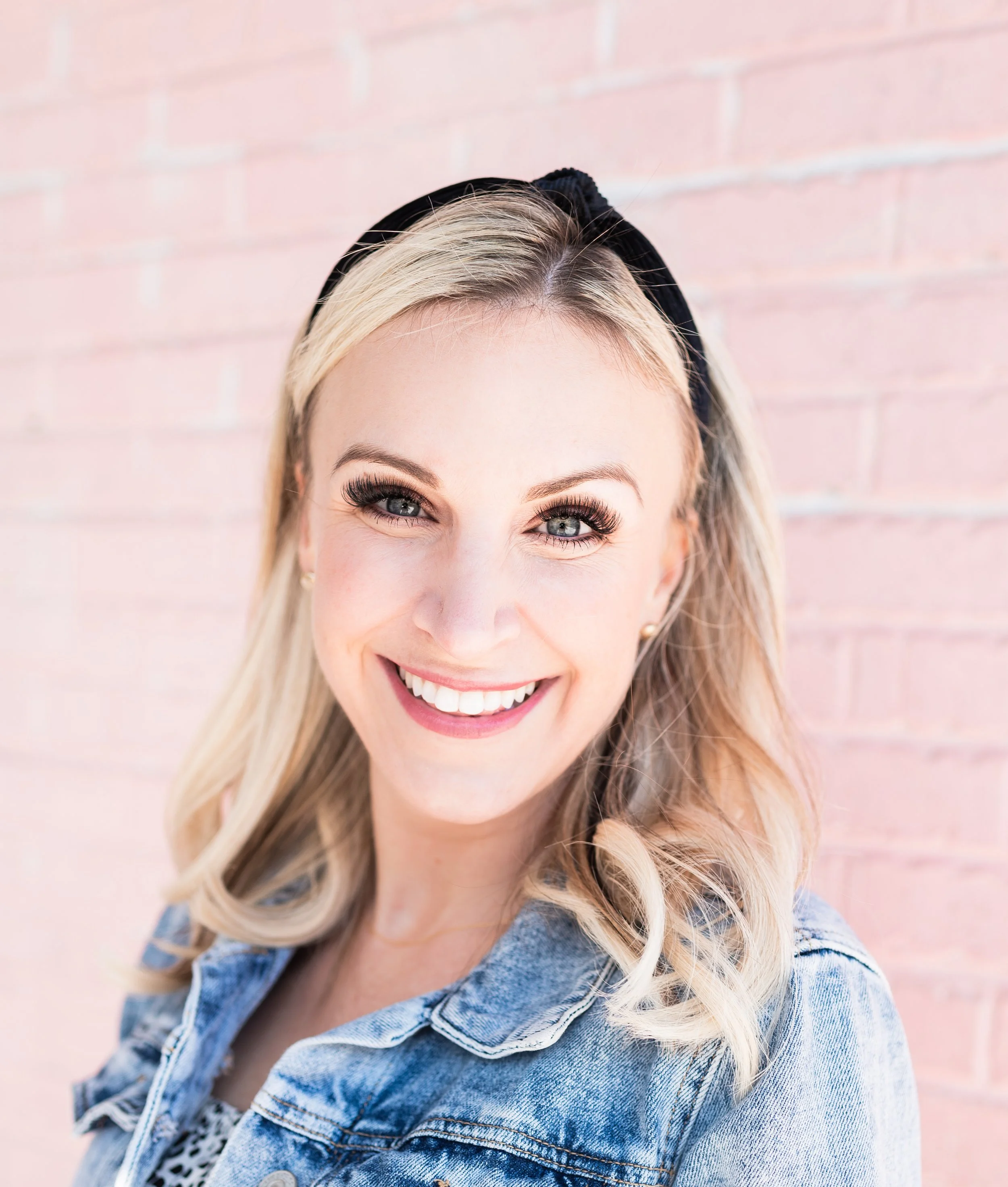 Close-up of a smiling woman with blonde hair, wearing a denim jacket and a black headband, standing in front of a pink brick wall.