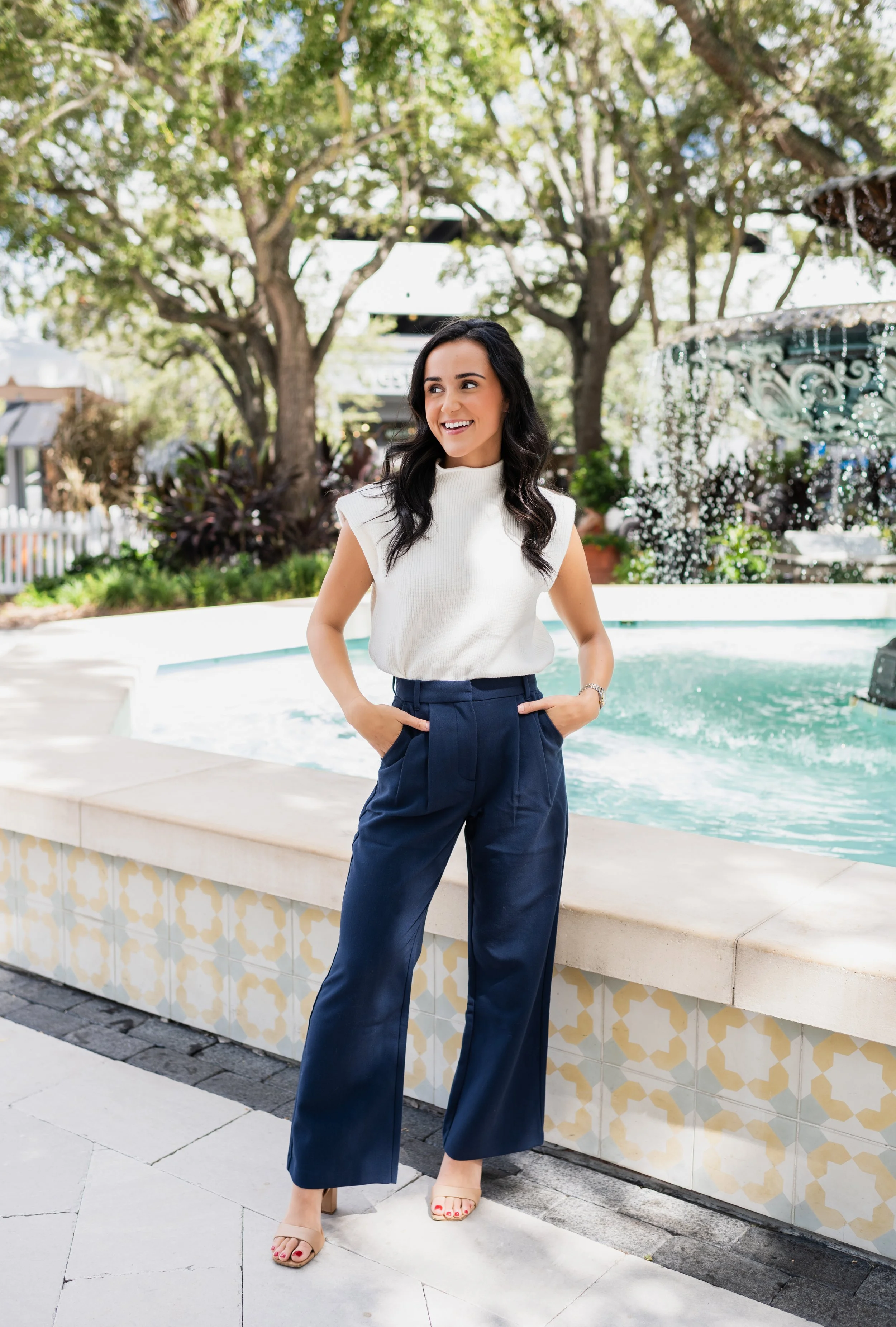 A woman standing outdoors by a fountain, smiling, wearing a white sleeveless top and dark blue wide-leg pants, with trees and a decorative fountain in the background.