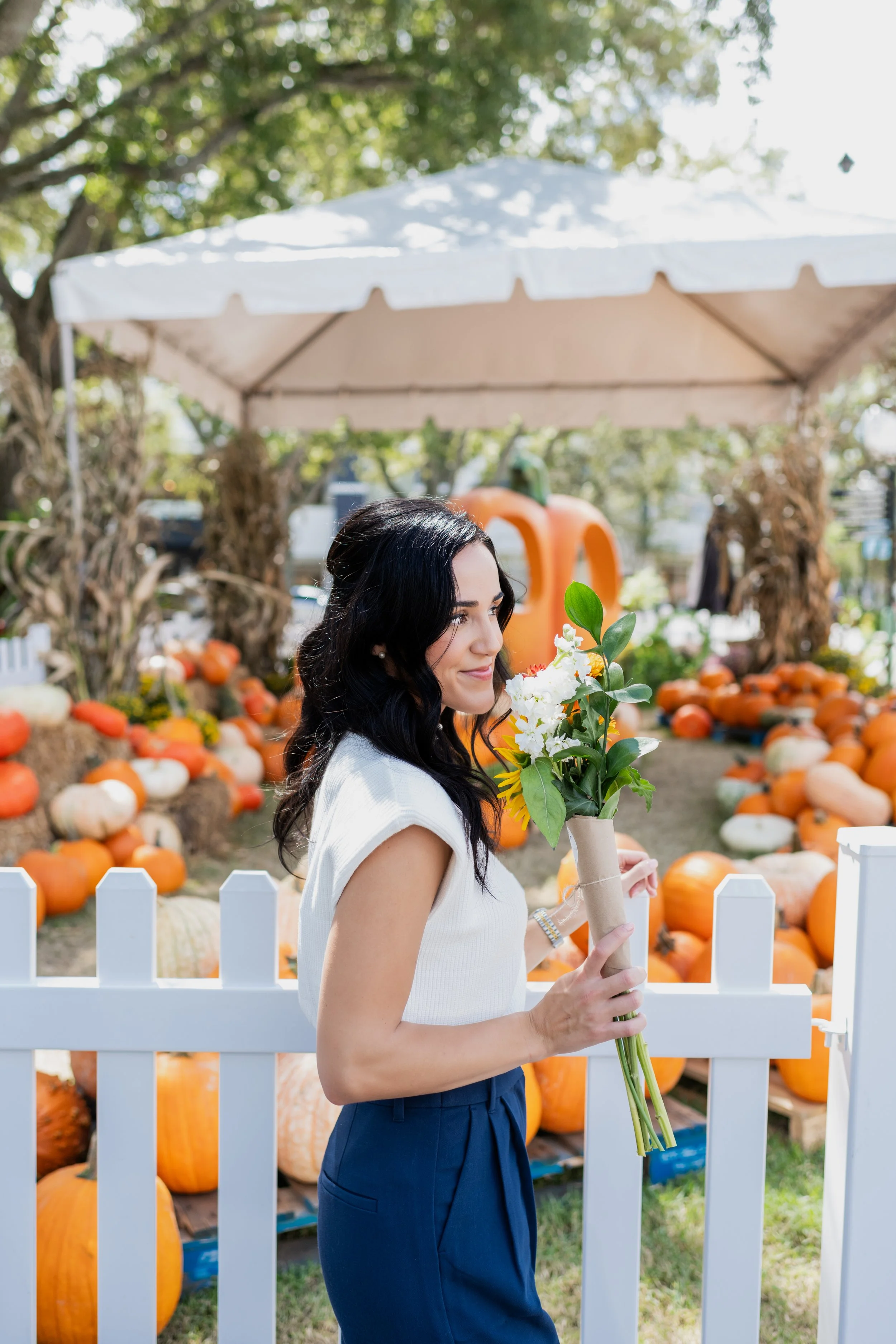 A woman holding a bouquet of white flowers and green leaves at a pumpkin patch, with pumpkins and hay bales in the background.
