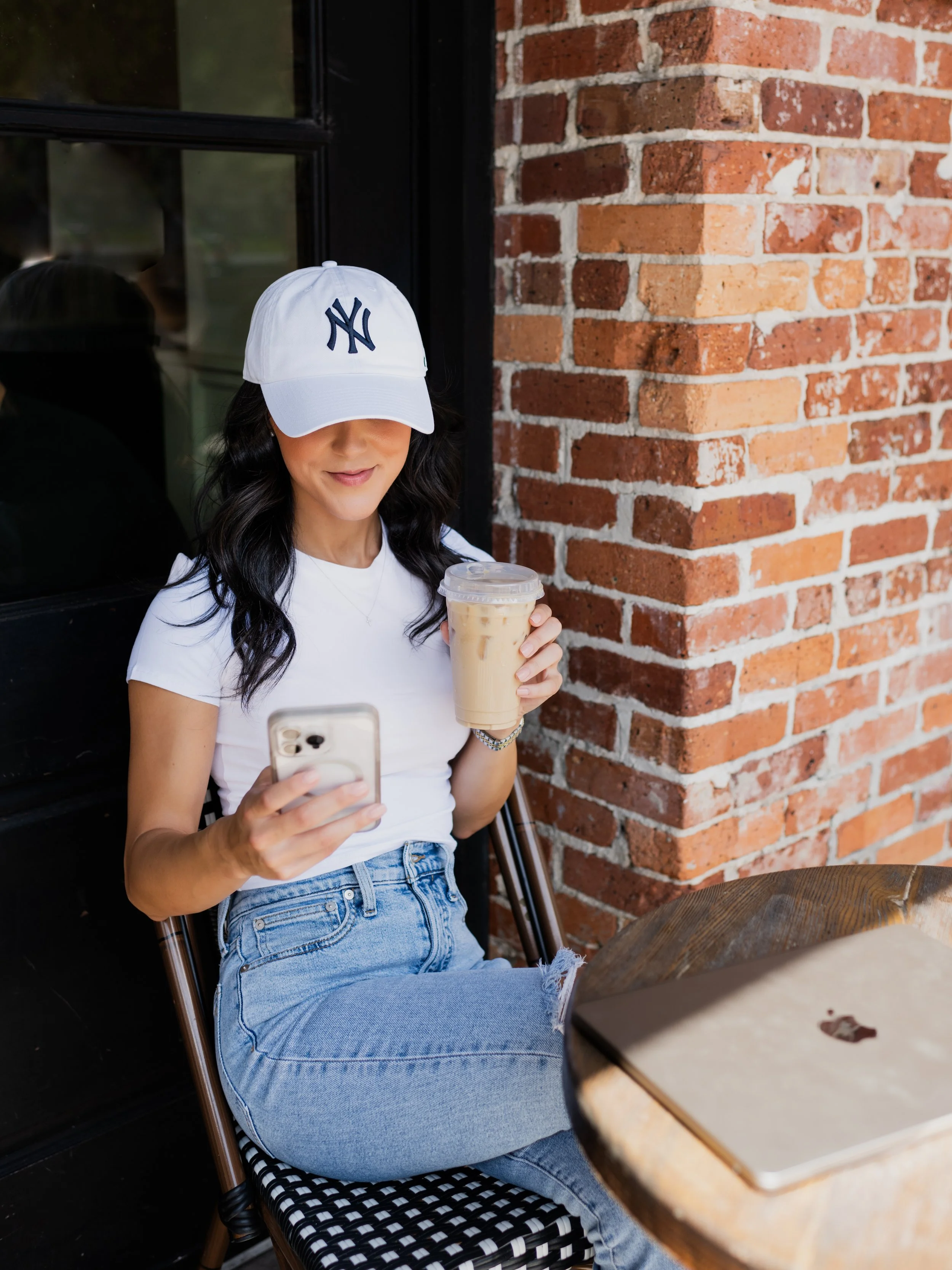A woman sitting outside at a cafe holds a cold coffee drink and a smartphone, wearing a white baseball cap with the New York Yankees logo, a white t-shirt, and ripped blue jeans.