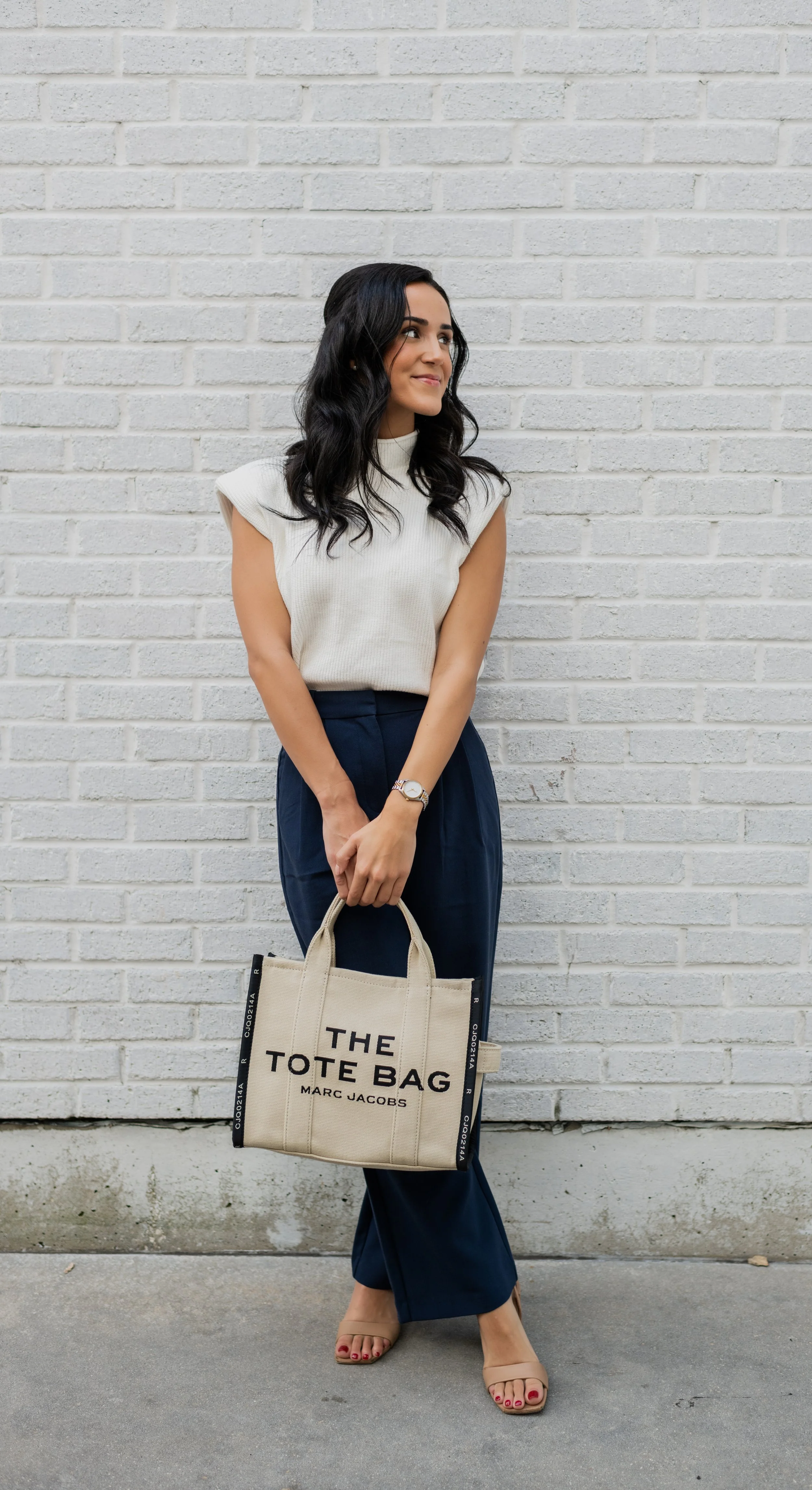 Woman in sleeveless white top and navy wide-leg pants standing against a white brick wall, holding a beige tote bag with black letters that read 'The Tote Bag - Marc Jacobs', wearing sand-colored open-toe heels.