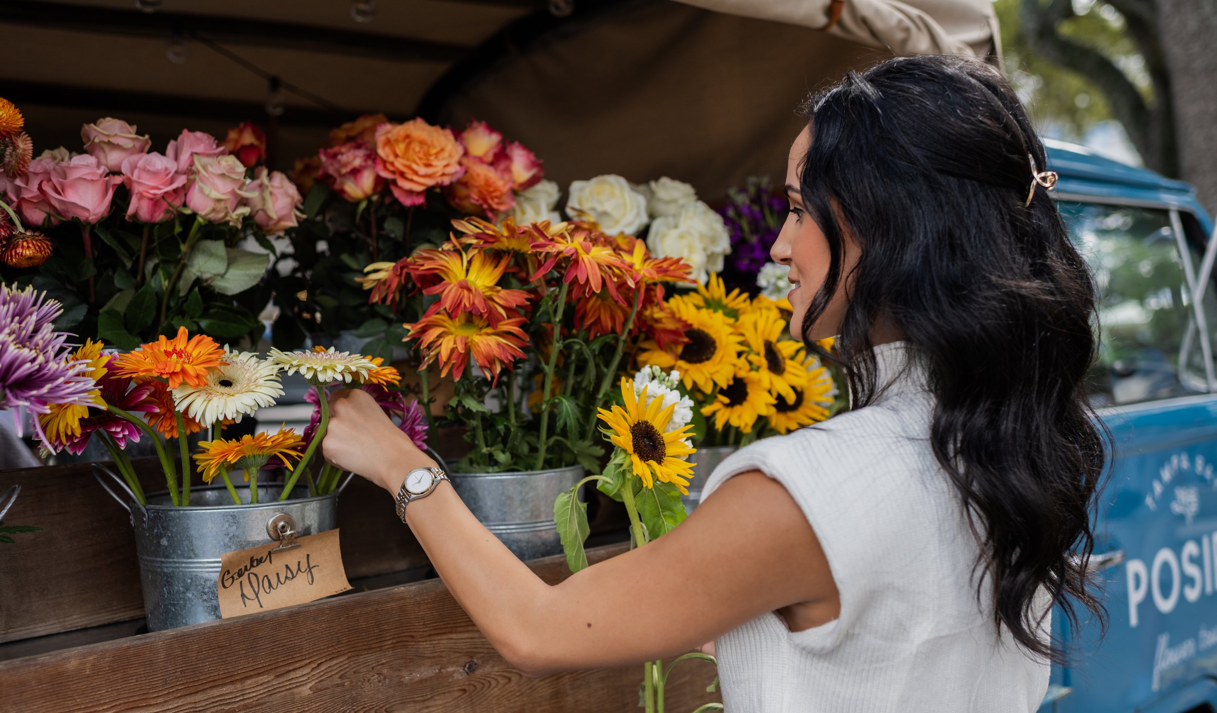 A woman with black wavy hair, wearing a white sleeveless top, shopping for flowers at a flower stand with buckets of colorful flowers, including daisies, sunflowers, and roses.