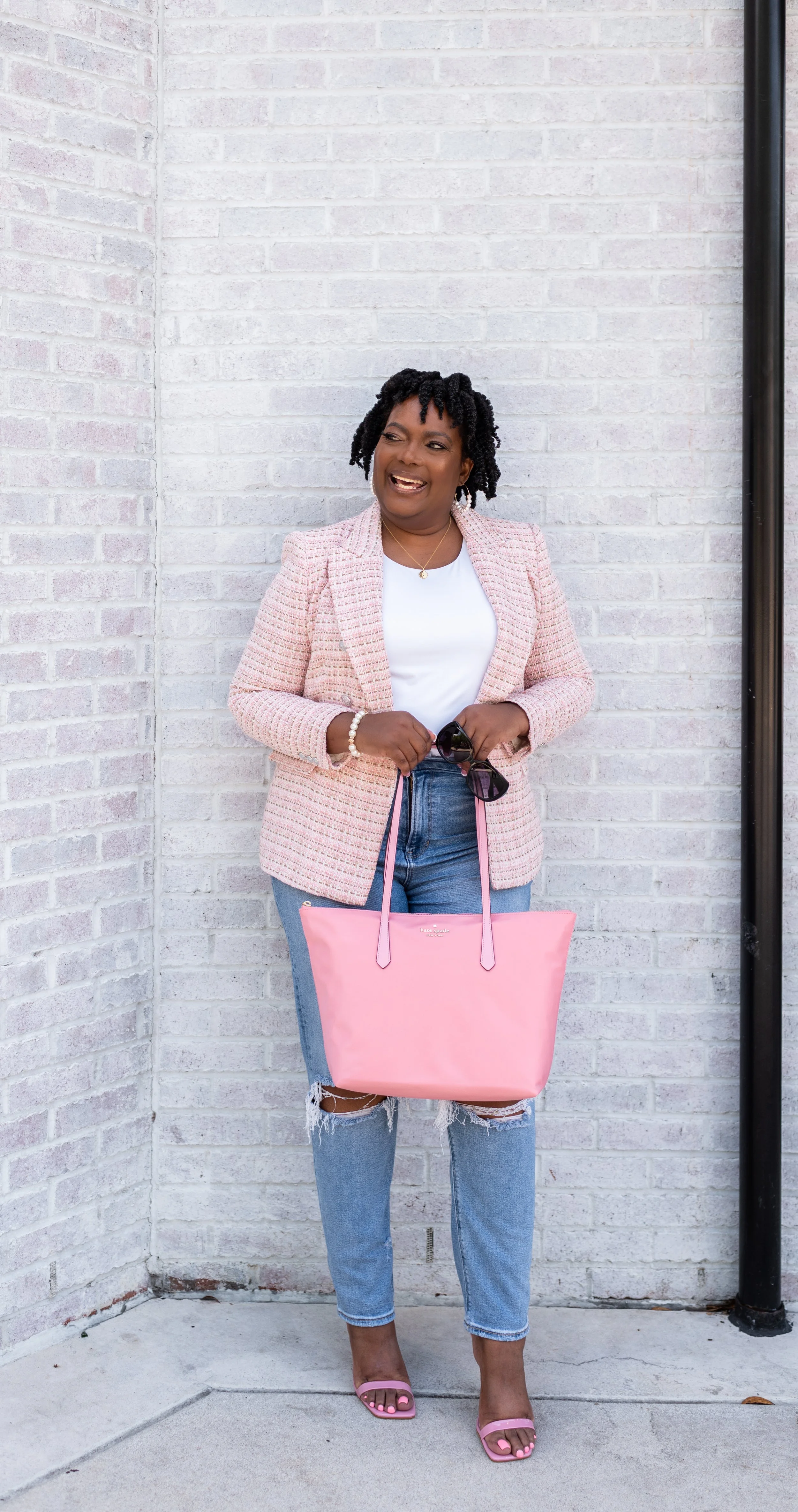 A woman standing against a white brick wall, holding sunglasses and a pink tote bag, wearing a pink blazer, white top, ripped jeans, and pink high-heeled sandals, smiling and looking to her right.