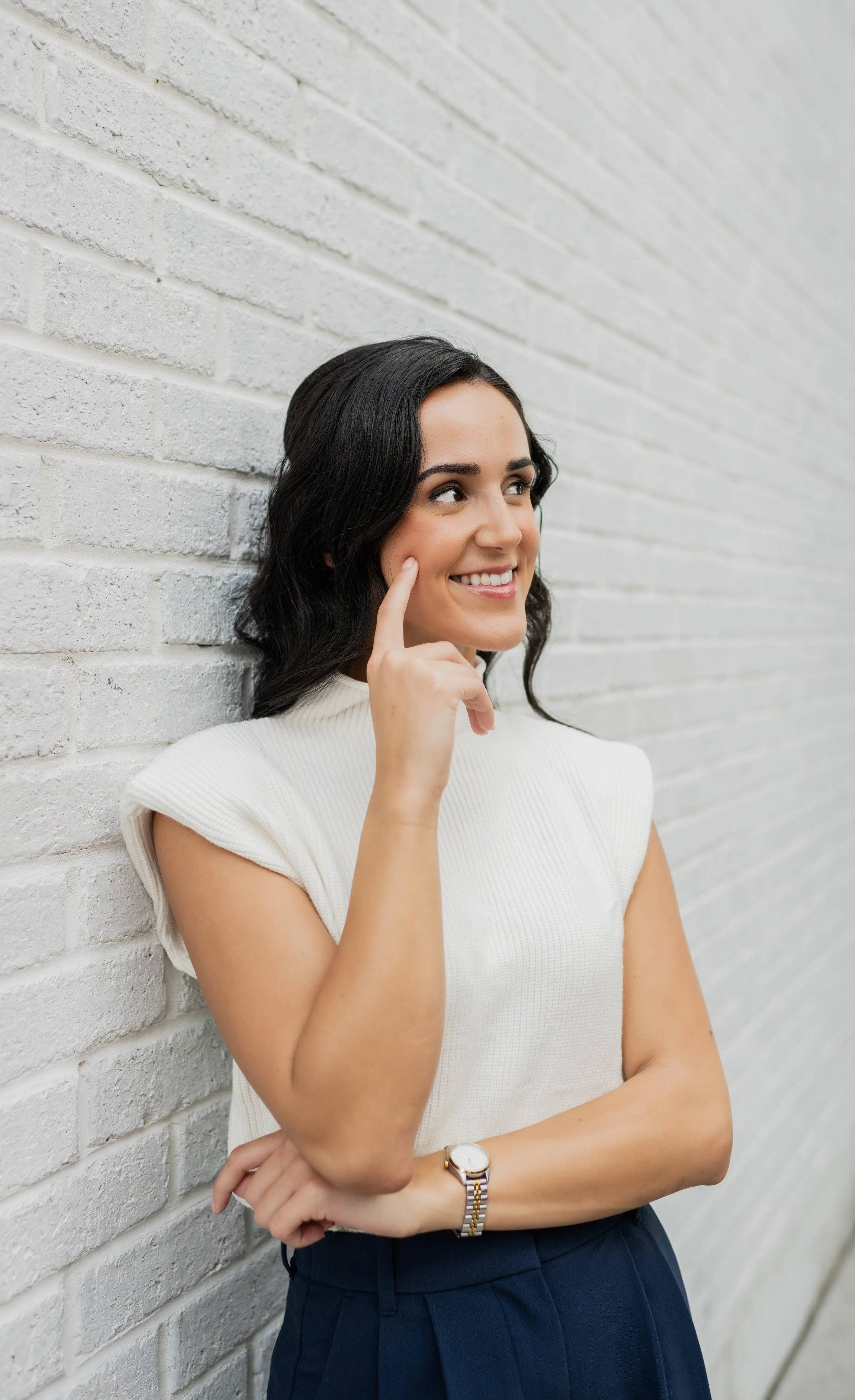 A young woman with black hair, wearing a white top and dark blue pants, standing against a white brick wall, smiling, and touching her cheek with her finger.