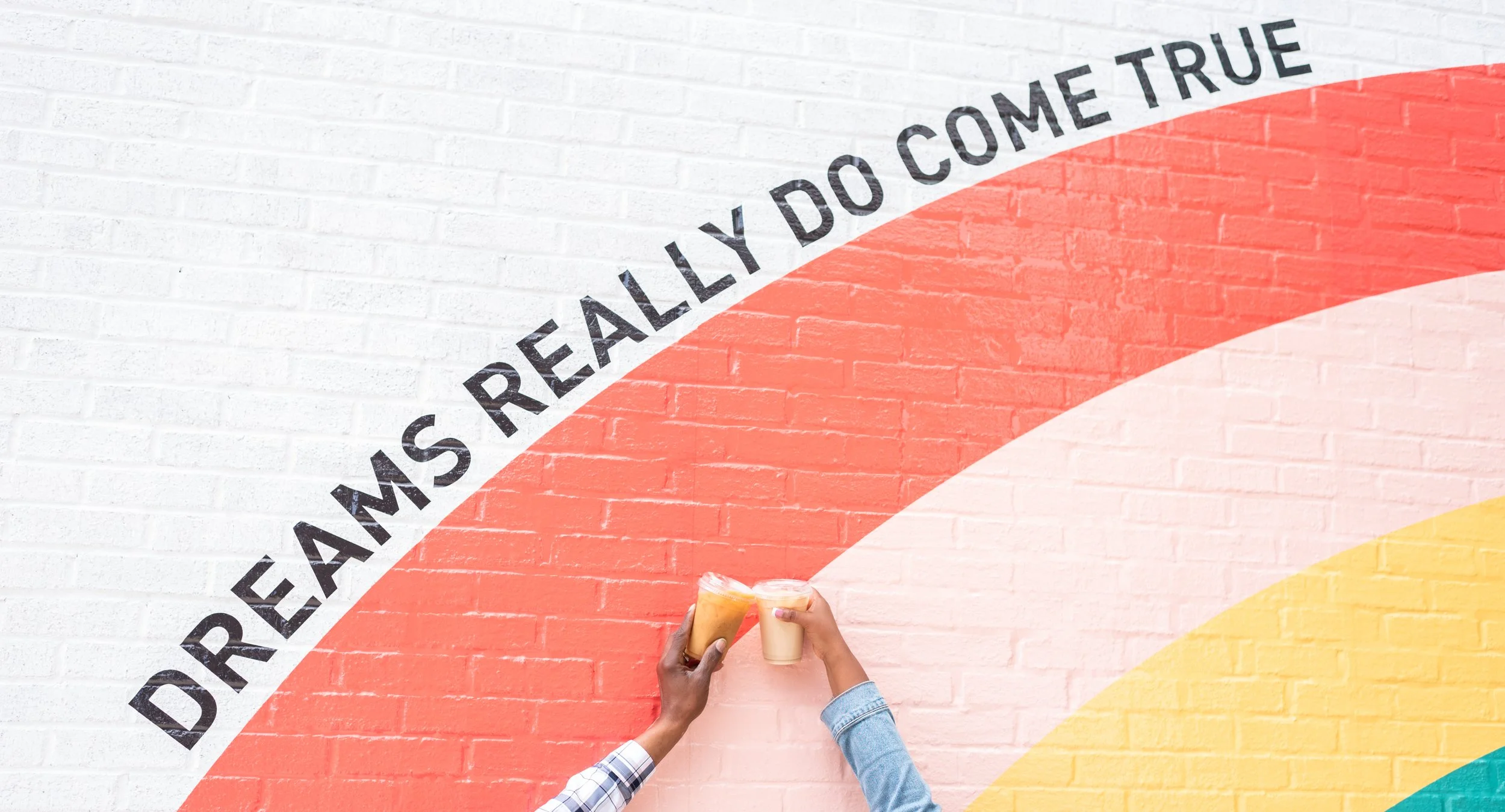 Two people holding plastic cups of drinks in front of a painted brick wall with rainbow-colored arcs and the phrase 'Dreams really do come true' written along the curves.