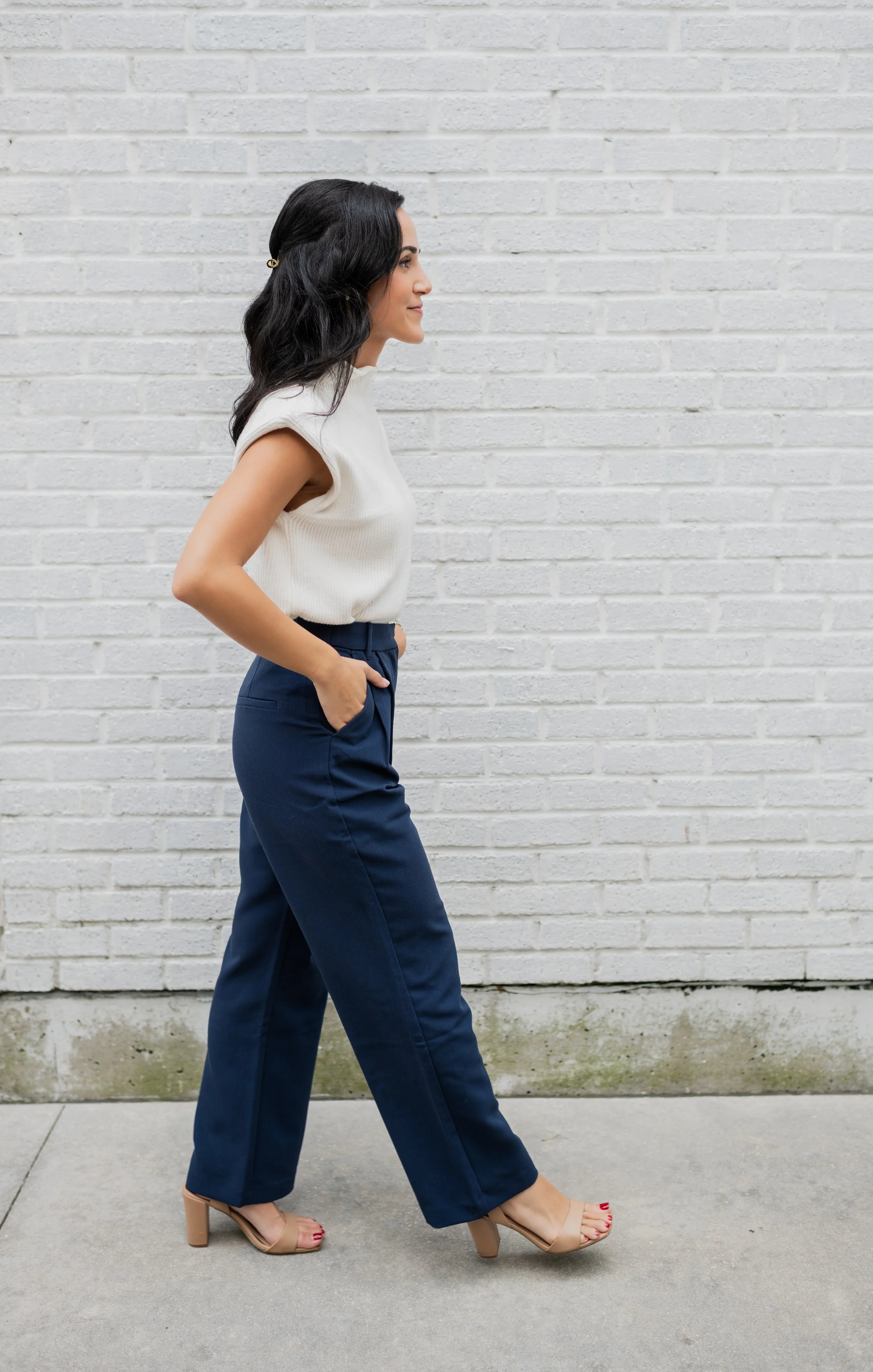 Side view of a woman with dark wavy hair in business casual attire, walking on a sidewalk against a white brick wall.