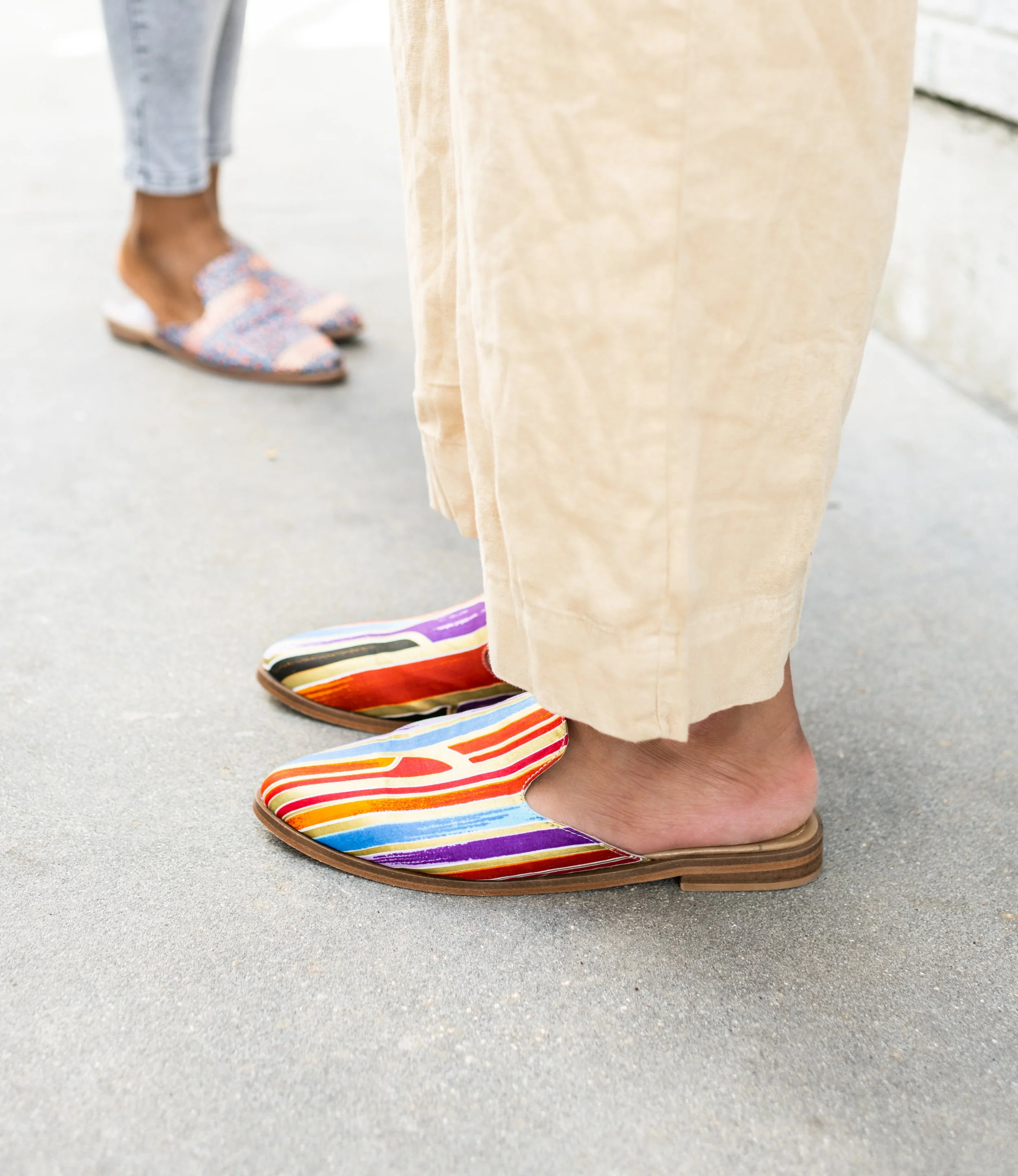 Person wearing colorful striped slip-on shoes standing on concrete sidewalk with another person in the background wearing patterned sneakers and gray pants.