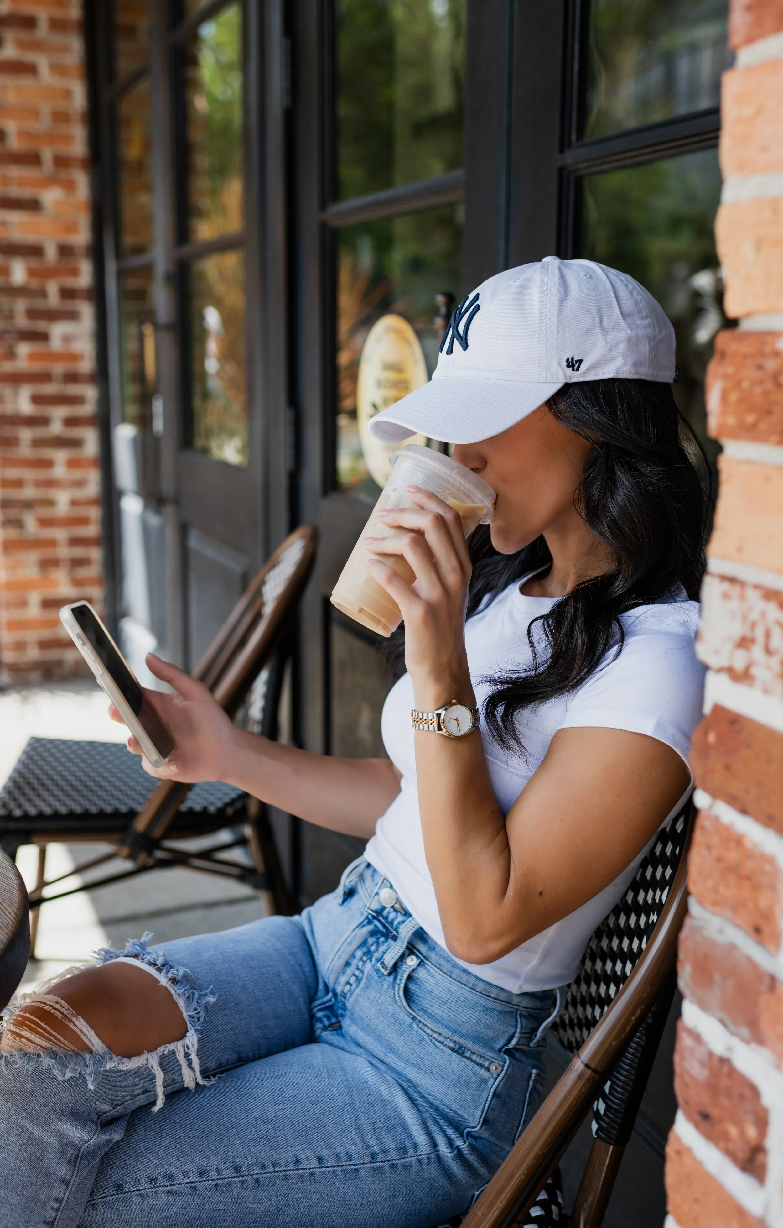 A woman sitting outside at a cafe, wearing a white baseball cap, white T-shirt, and ripped jeans, sipping iced coffee from a plastic cup while looking at her smartphone.